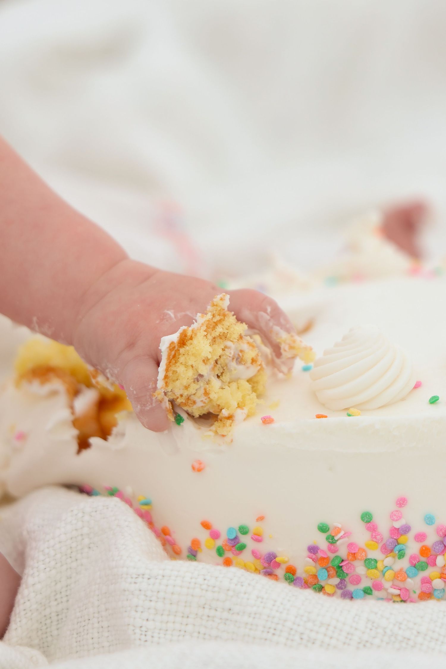 A small hand grabs cake with white frosting and colorful sprinkles against a soft textured fabric background.