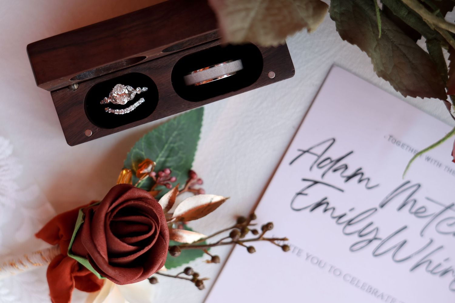 Wooden ring box next to a wedding invitation  and red rose against a white surface.
