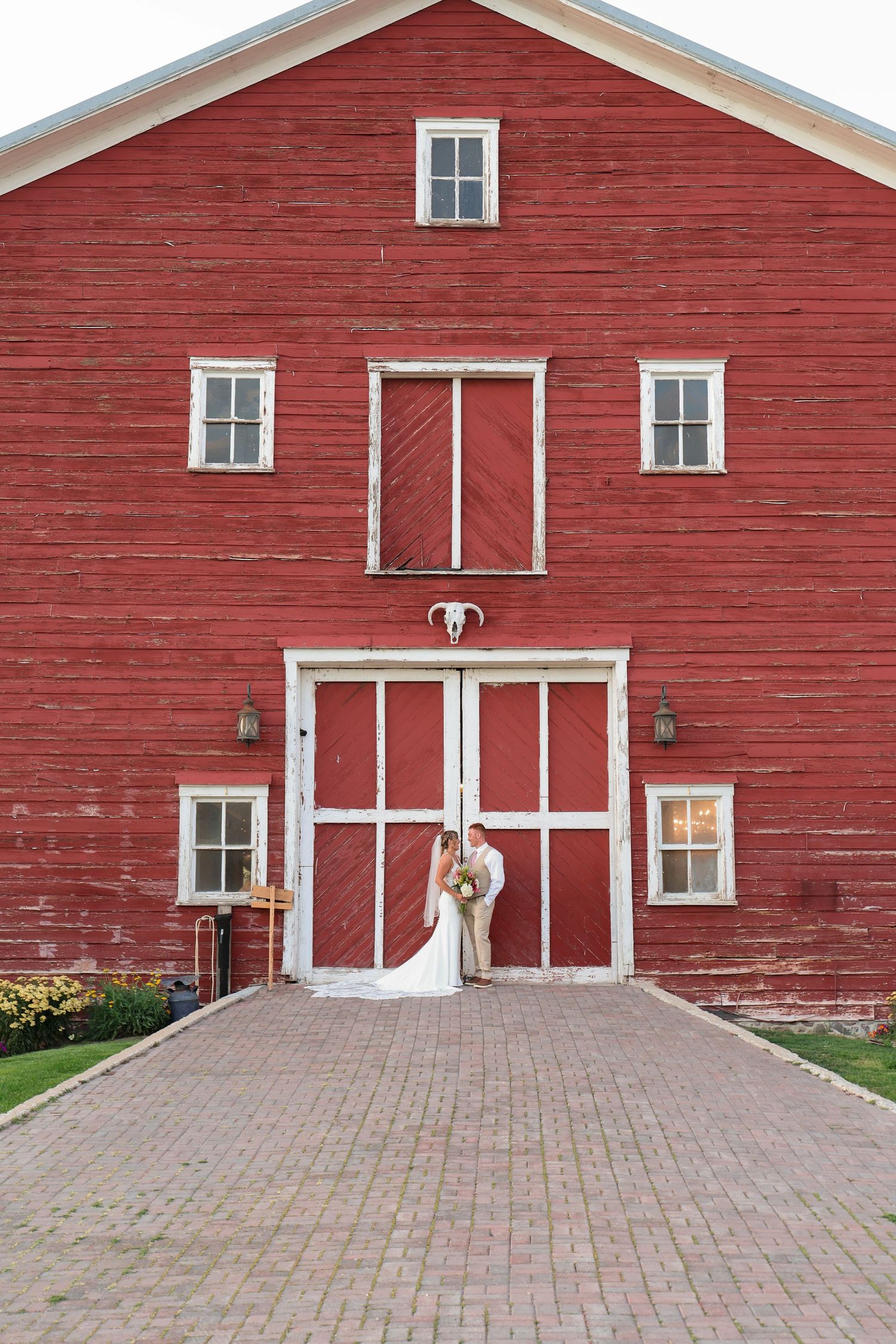 A bride and groom pose in front of  classic red barn with white trim features, traditional barn doors and a brick paver walkway in front.