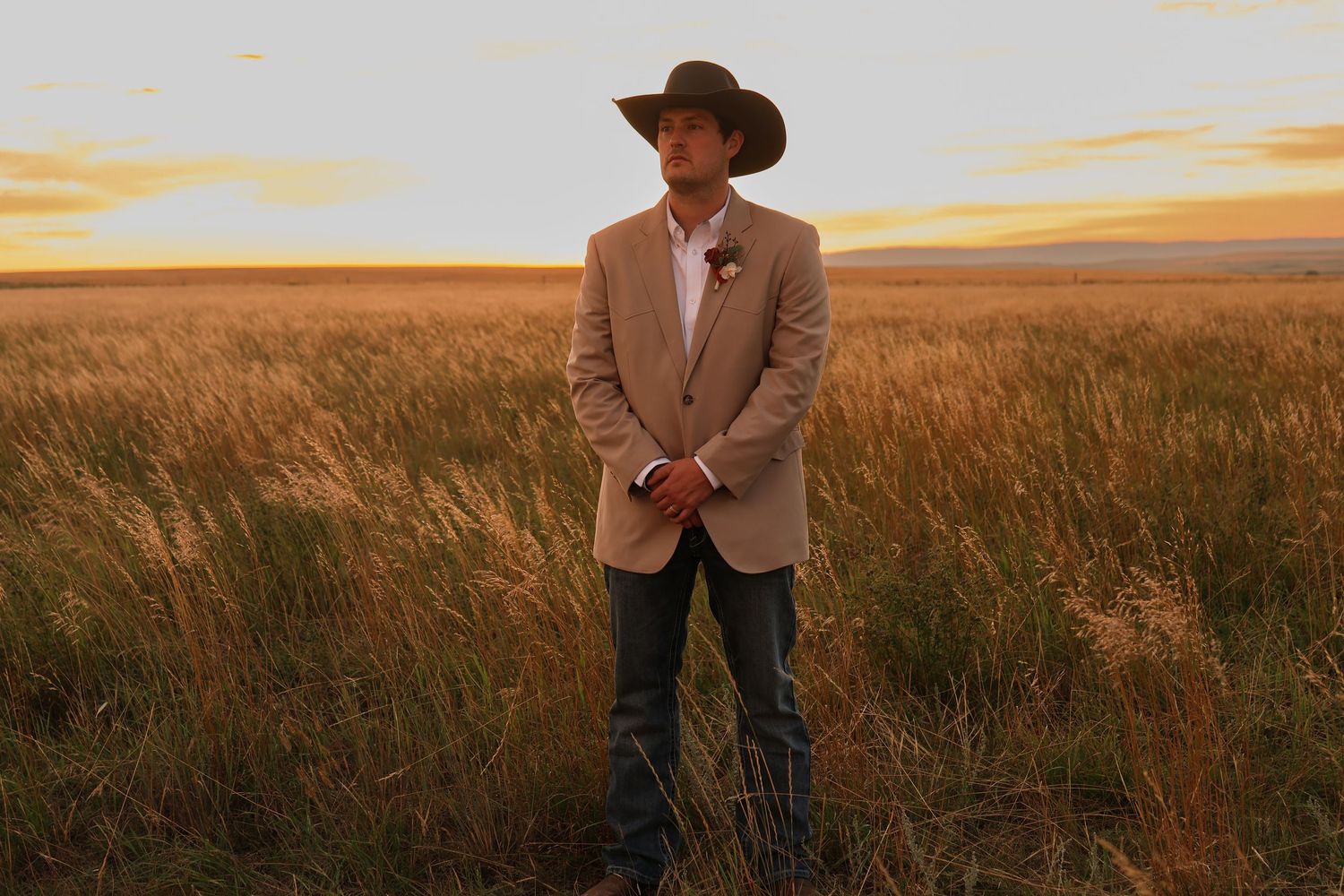 A groom in a cowboy hat stands in golden prairie grass at sunset amid golden glowing light.