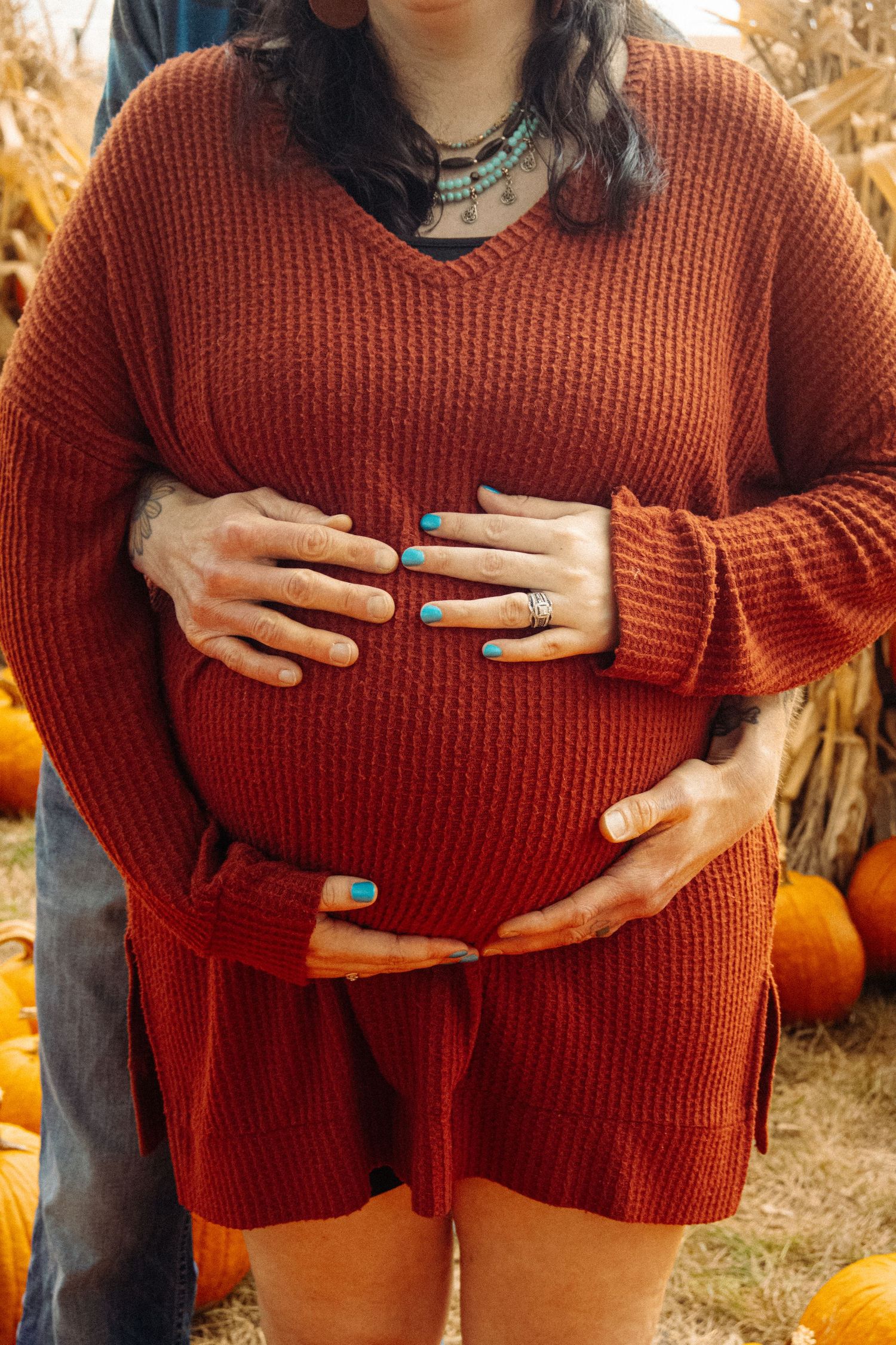 Person in red sweater touching their belly while surrounded by pumpkins and corn stalks.