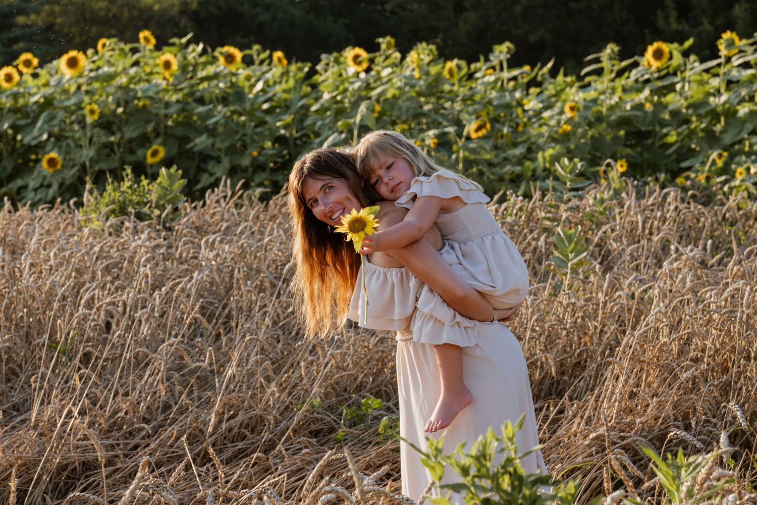 Woman in white dress holds smiling child near sunflower field on sunny day.