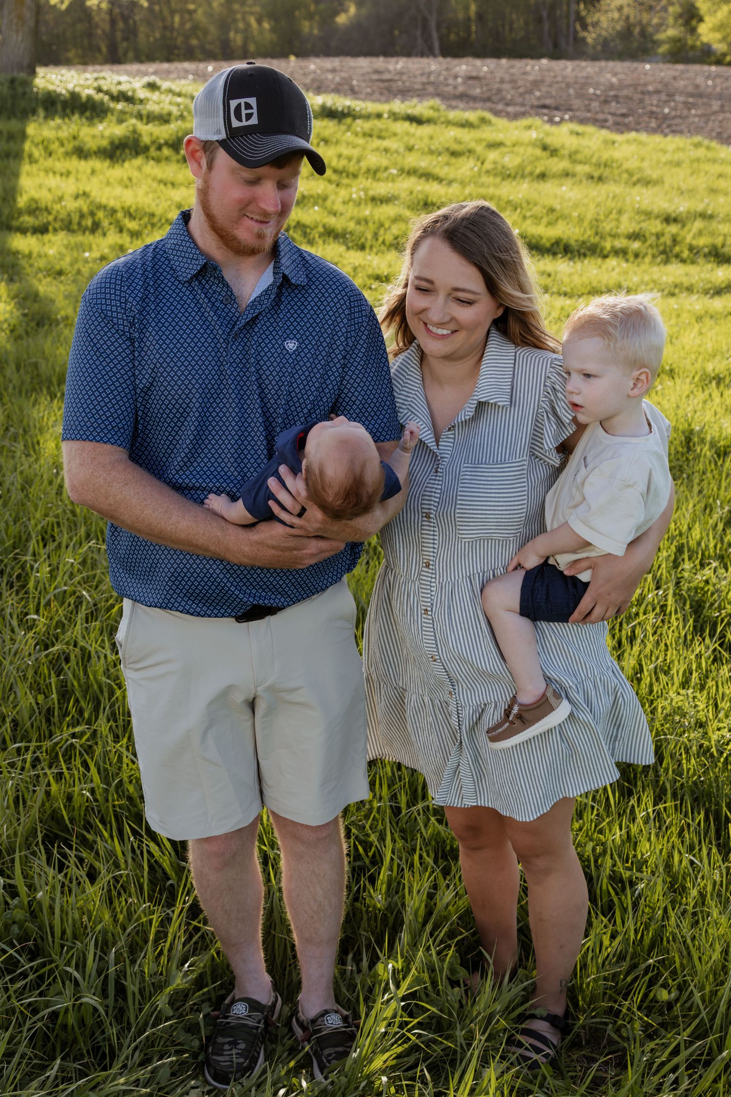 A happy young family stands in a green field, with dad holding a newborn and mom carrying a toddler on her hip.
