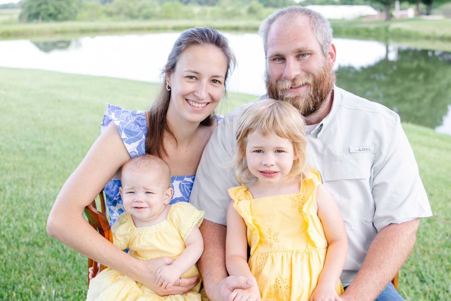 Family of four posing outdoors near a pond, two parents with baby and toddler in yellow dresses on green grass.