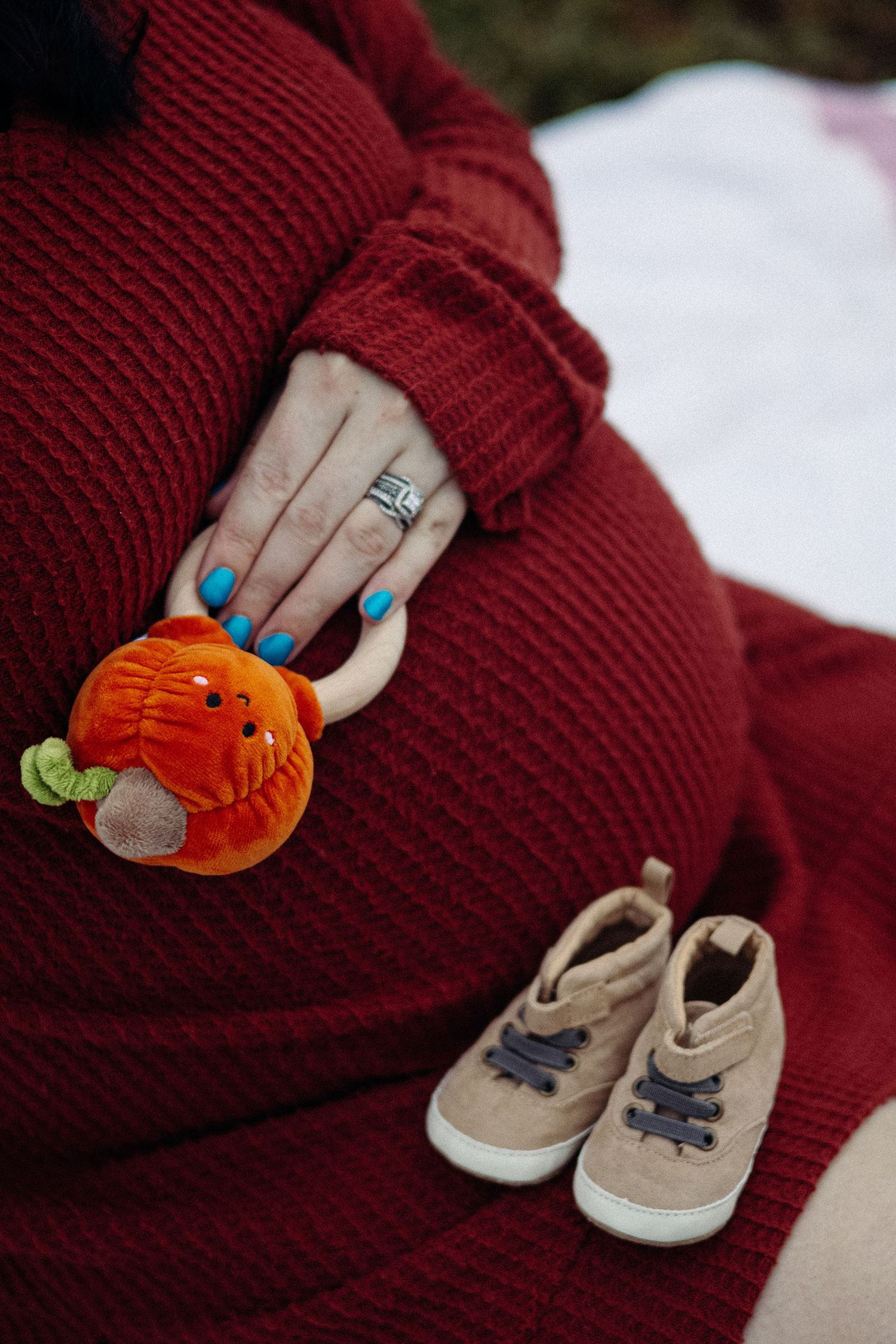 A pair of small beige baby shoes next to a pumpkin and hands with blue nail polish.