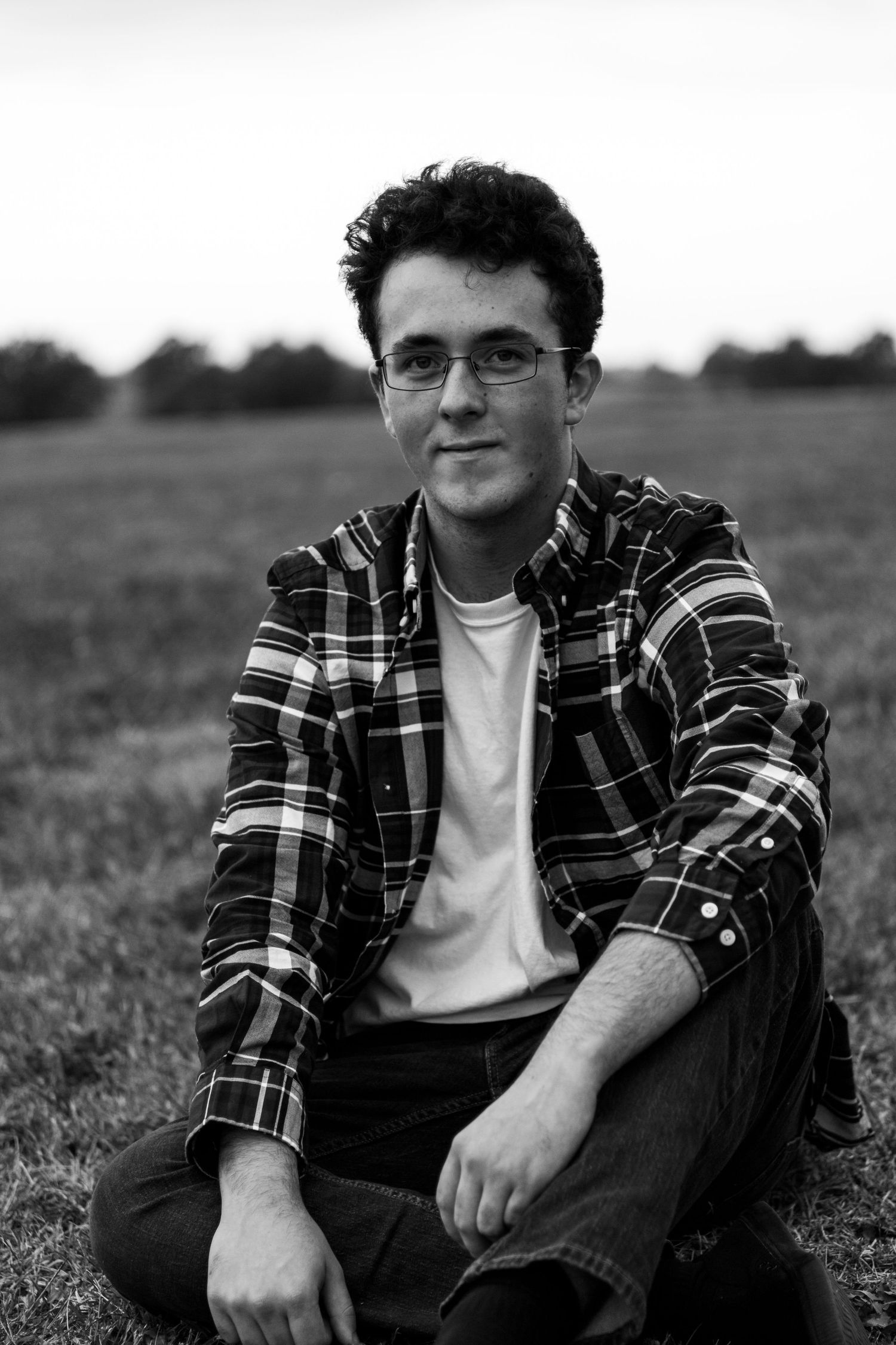 Black and white portrait of person sitting in grass wearing plaid shirt and glasses.