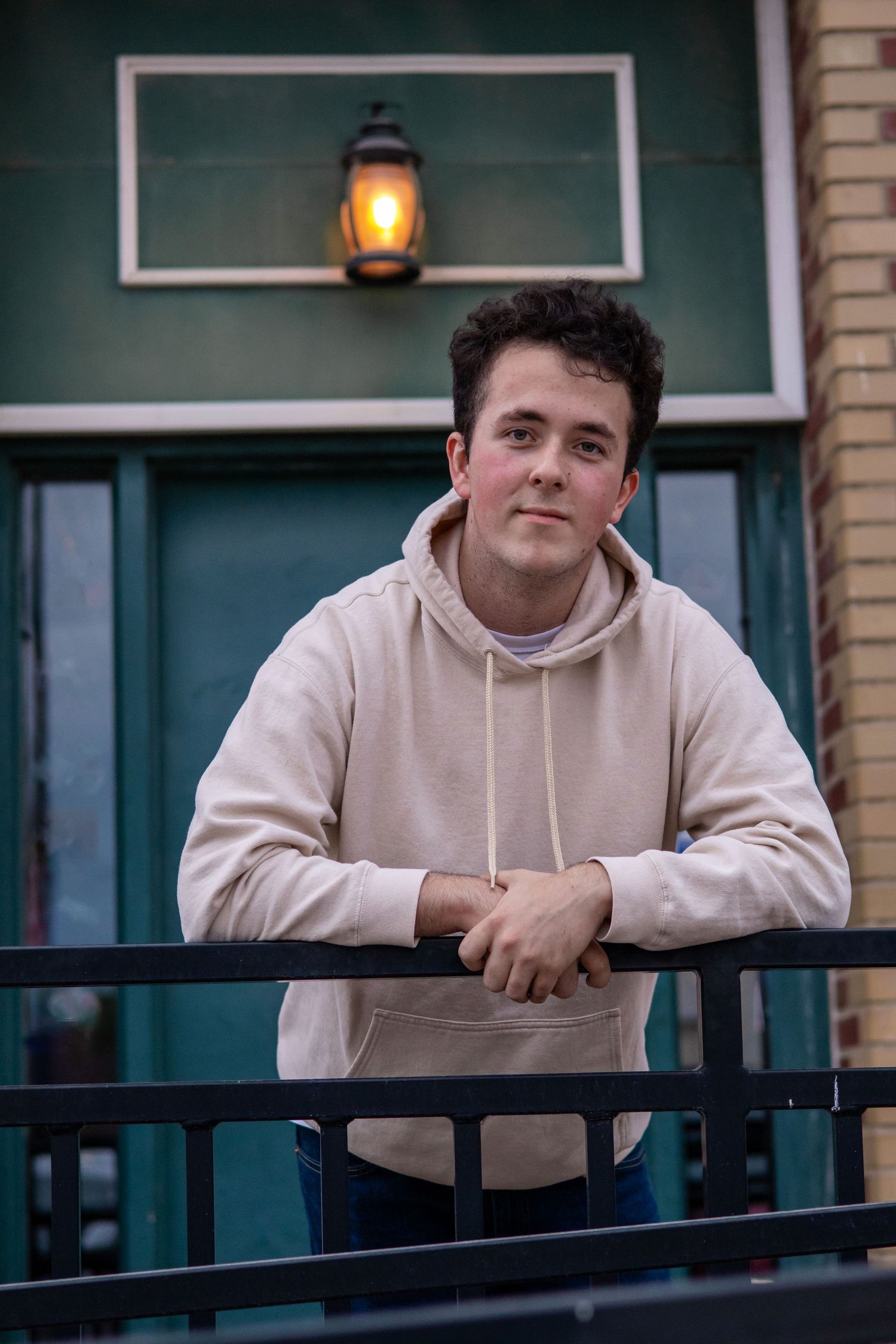 Person in beige hoodie leaning on black railing with green building exterior in background.