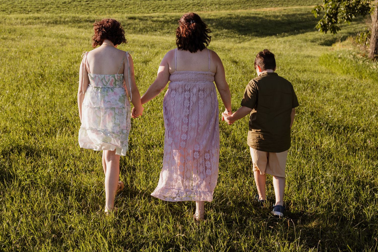 Three people walking hand-in-hand through a green field, two in dresses and one in shorts, viewed from behind.