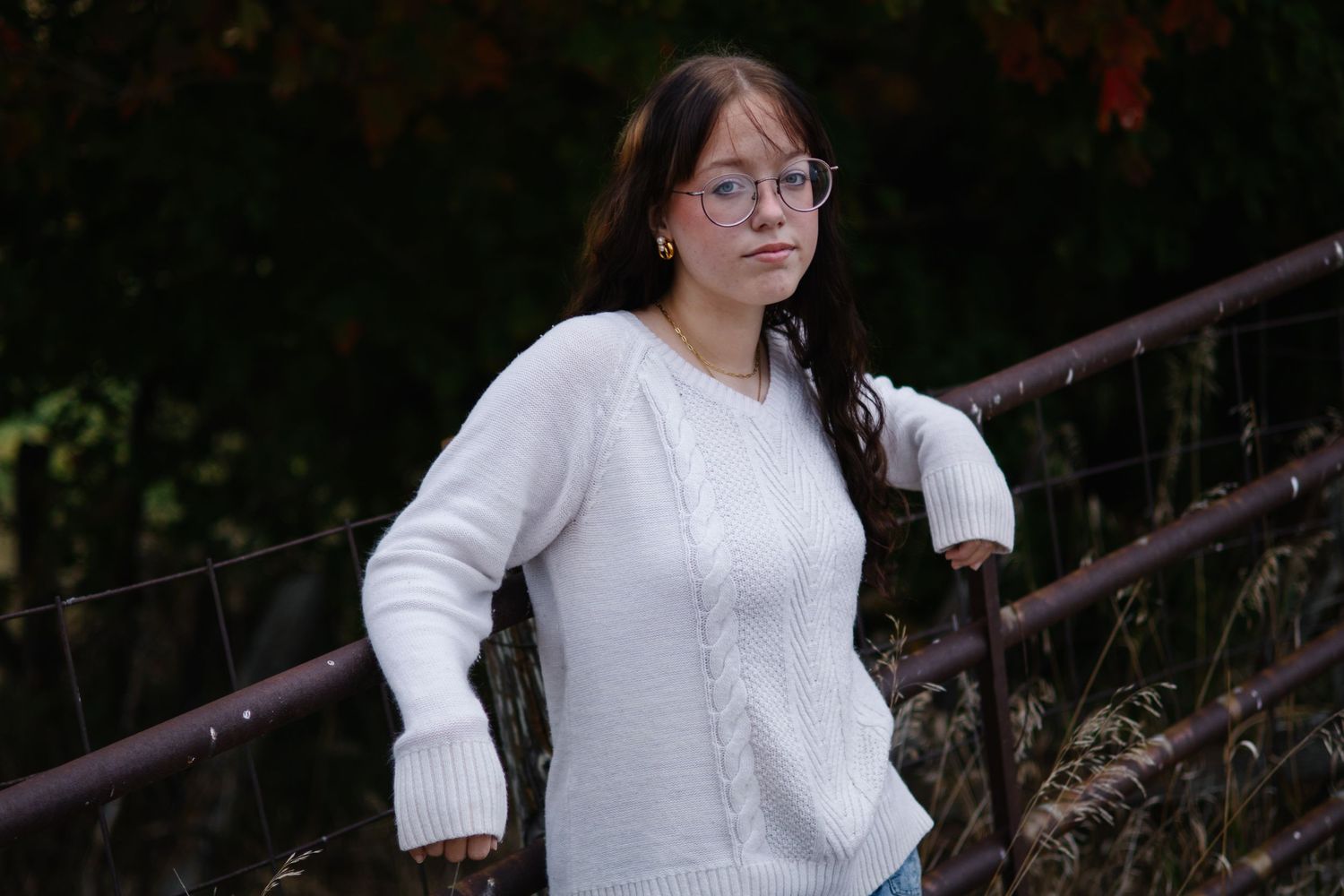 A person in a white sweater and glasses poses outdoors in a natural setting with trees and sunlight.