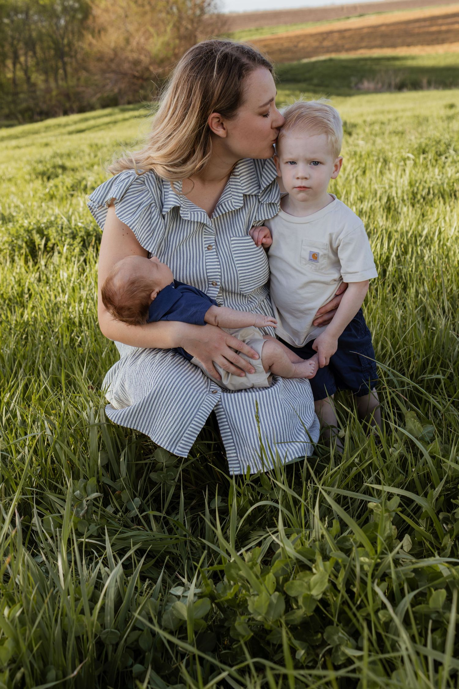 Mother kissing toddler son while holding newborn baby in lush green grass field during golden hour.