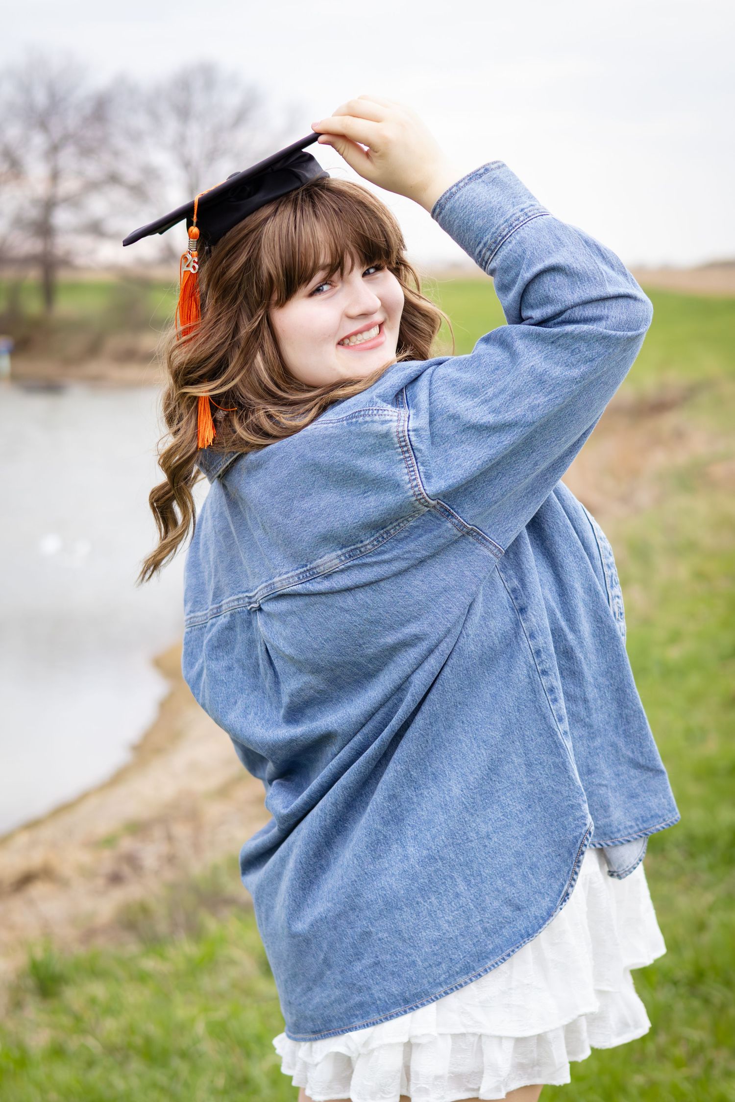 Smiling young woman in denim jacket and graduation cap poses outdoors near a lake celebrating her academic achievement.