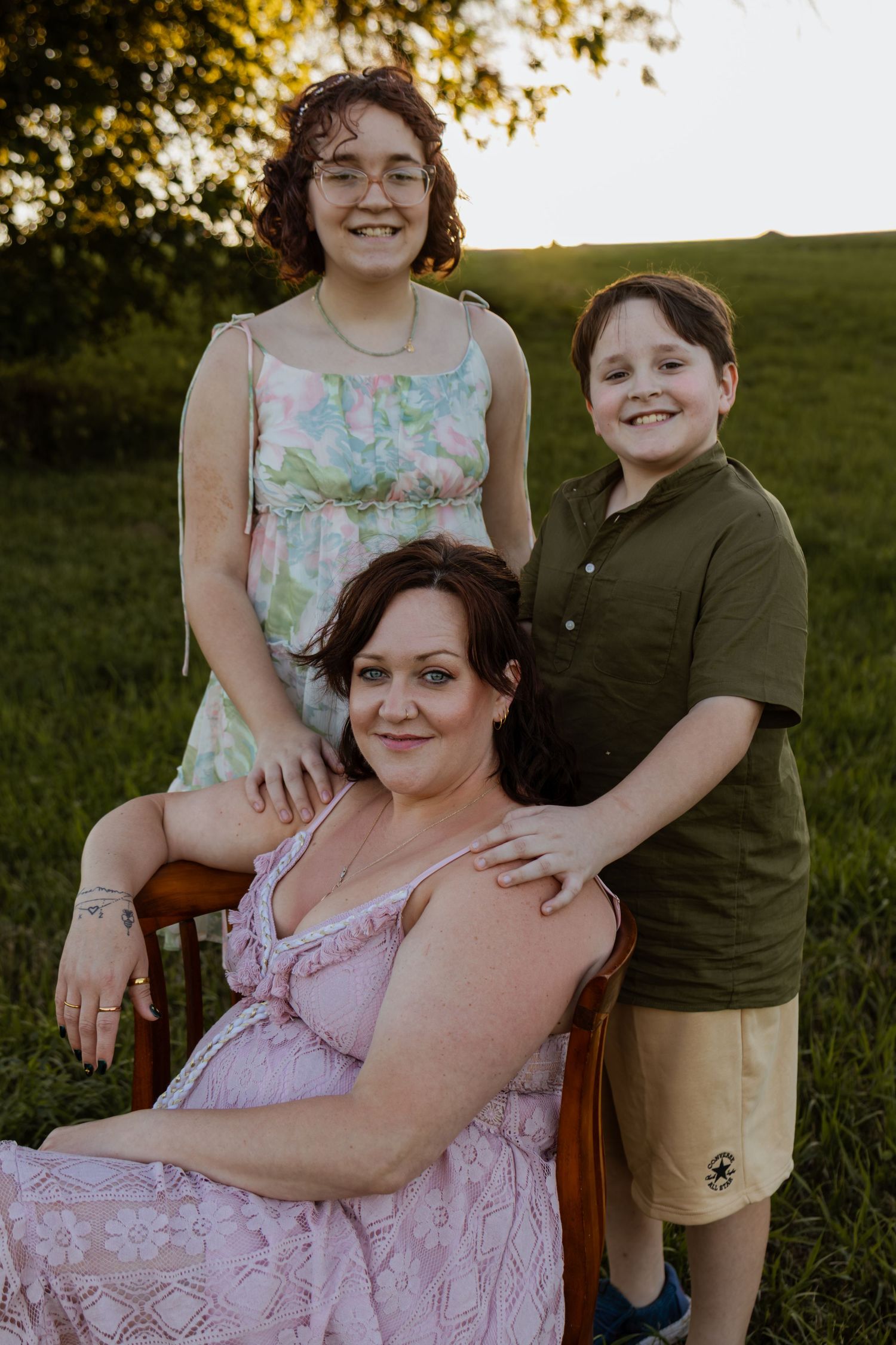 A woman in a pink dress sits in a chair outdoors, flanked by two smiling children, with a green field at sunset.