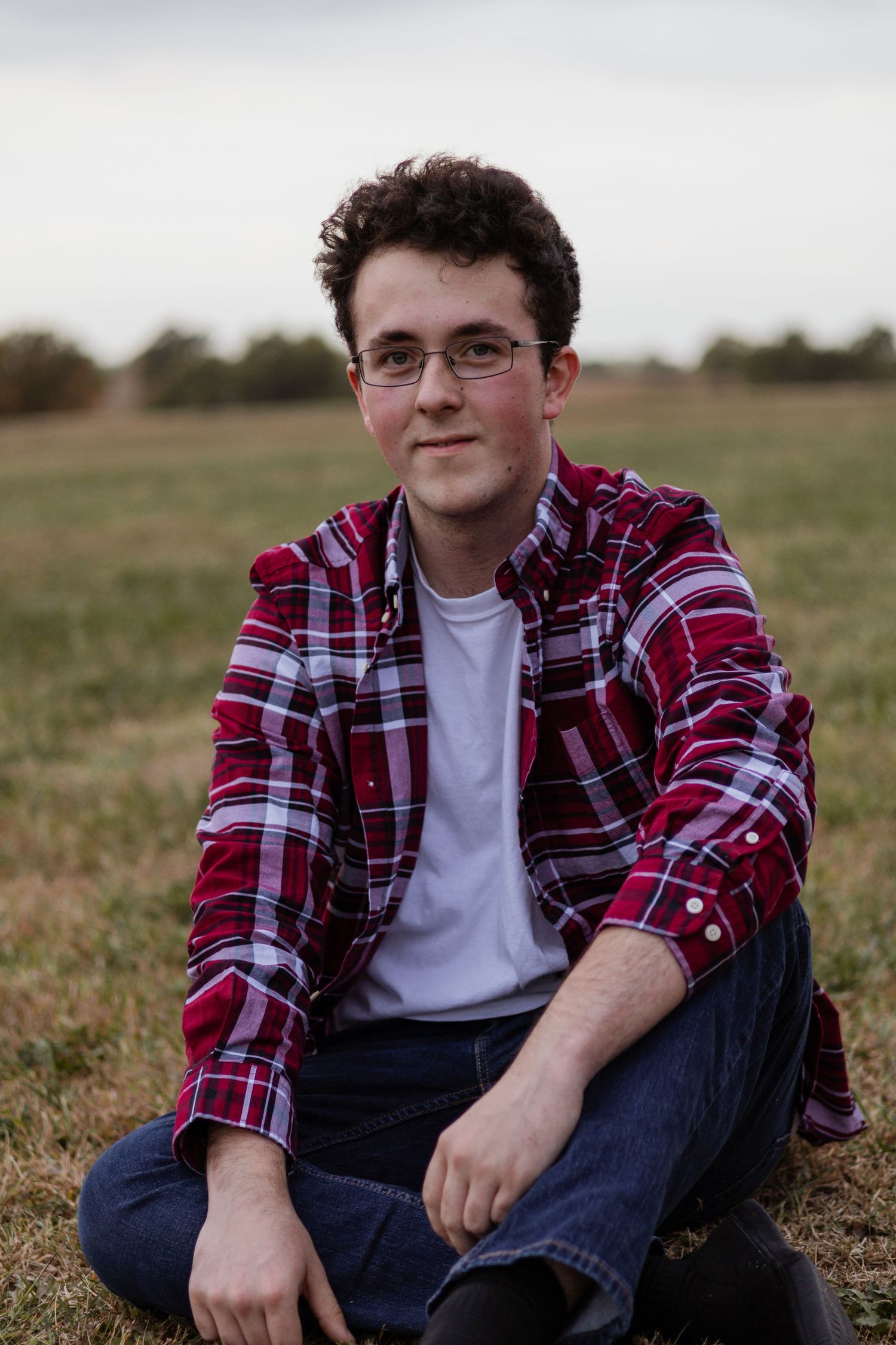 A person in a red plaid flannel shirt and glasses sits casually in a grassy field on an autumn day.