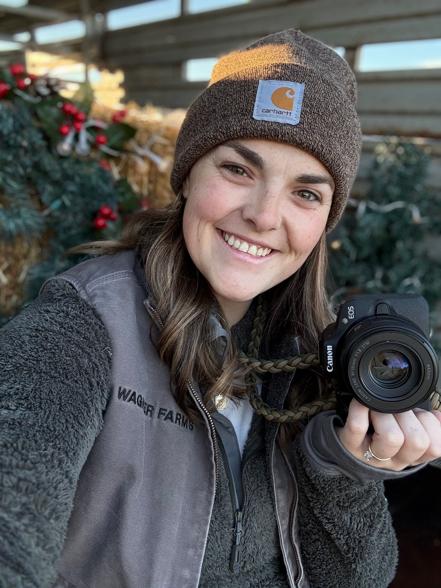 Smiling woman in Carhartt beanie and vest holds Canon camera outdoors near festive Christmas tree display.