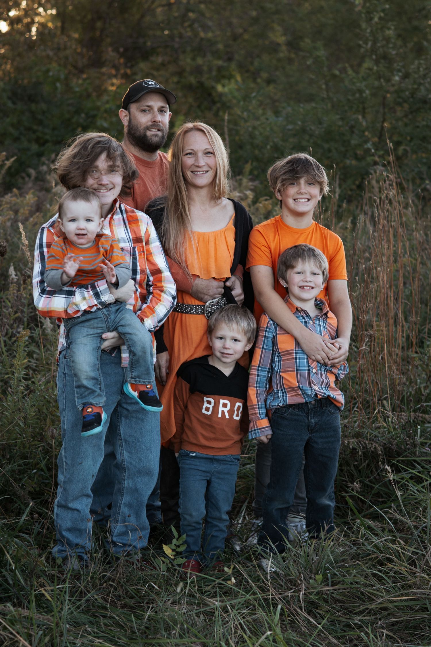 Family wearing matching orange shirts and overalls posing together in tall grass at sunset.
