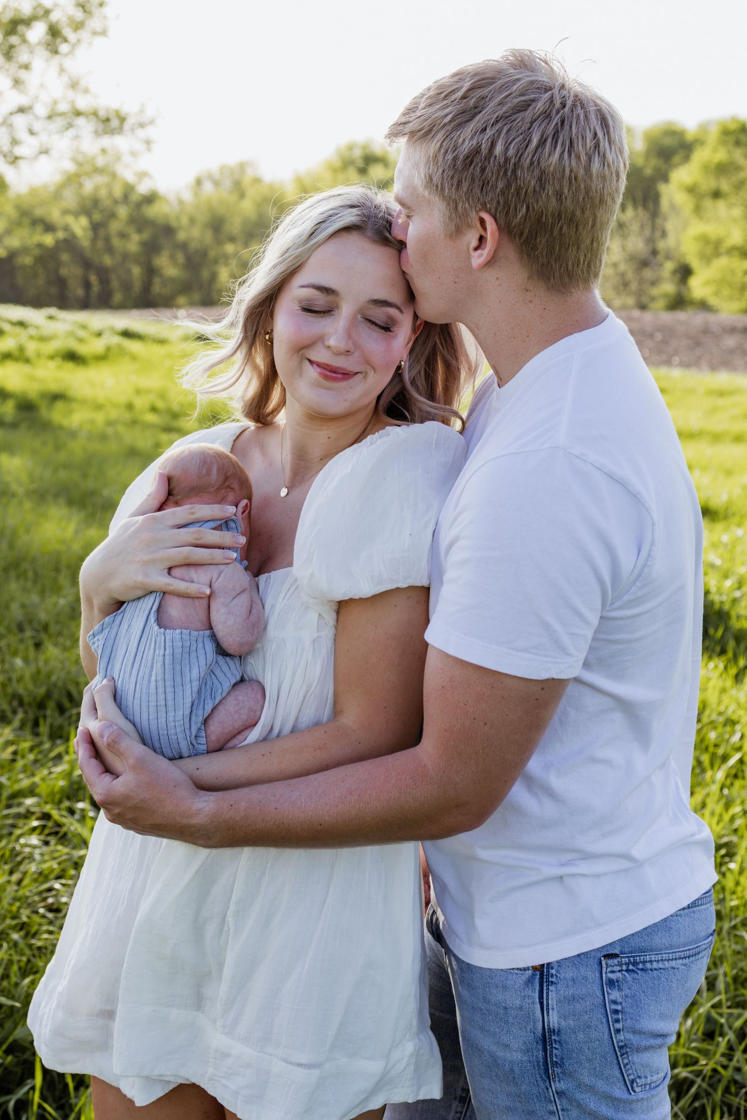 Young couple outdoors in green field, man kissing woman on cheek as she smiles holding newborn baby in blue wrap.