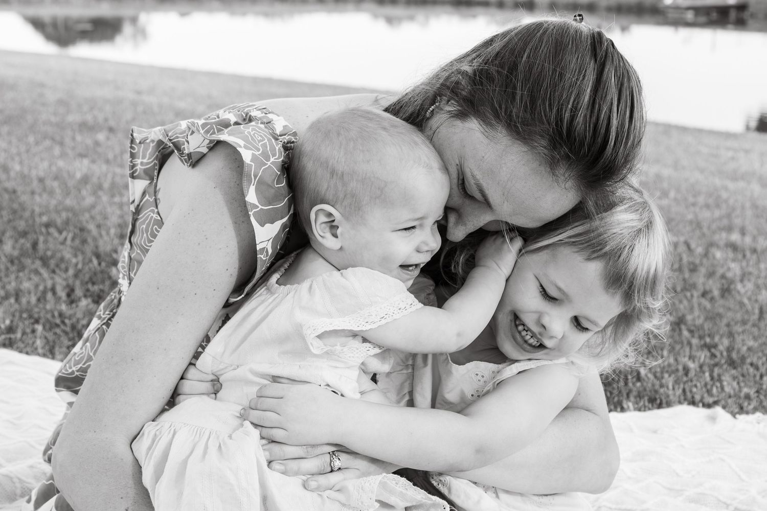 Black and white photo of a mother lovingly embracing her baby and young daughter outdoors near a lake.