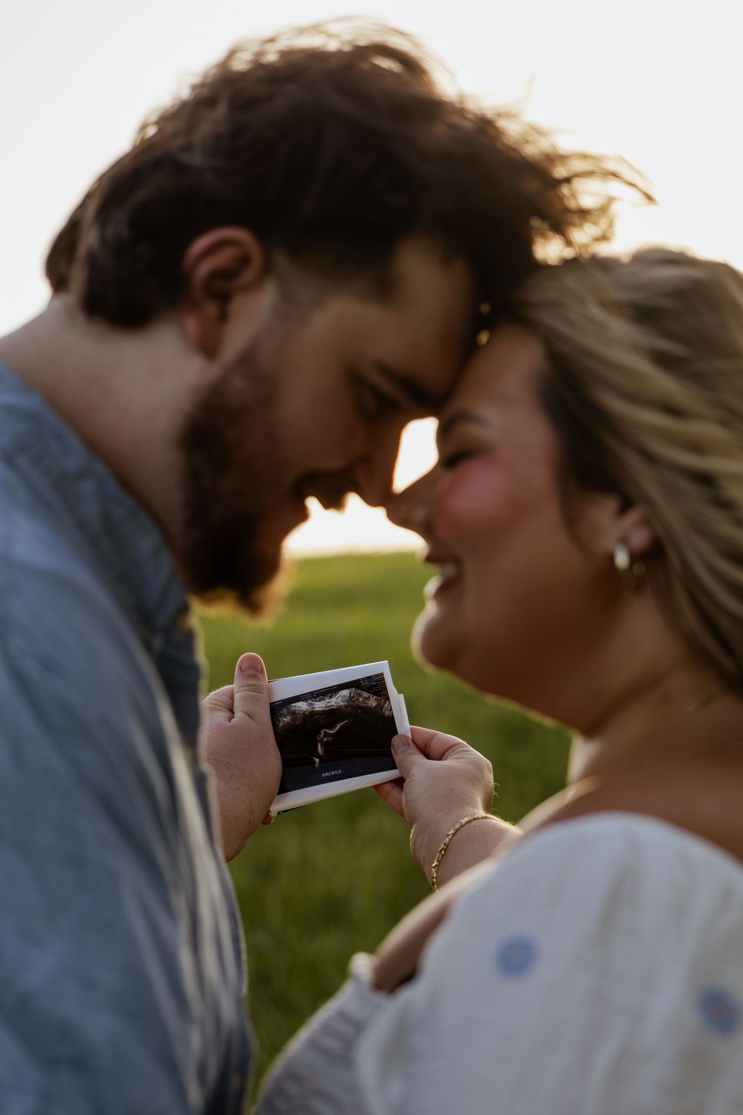 A joyful couple touching foreheads while holding an ultrasound photo outdoors in a green field at golden hour.