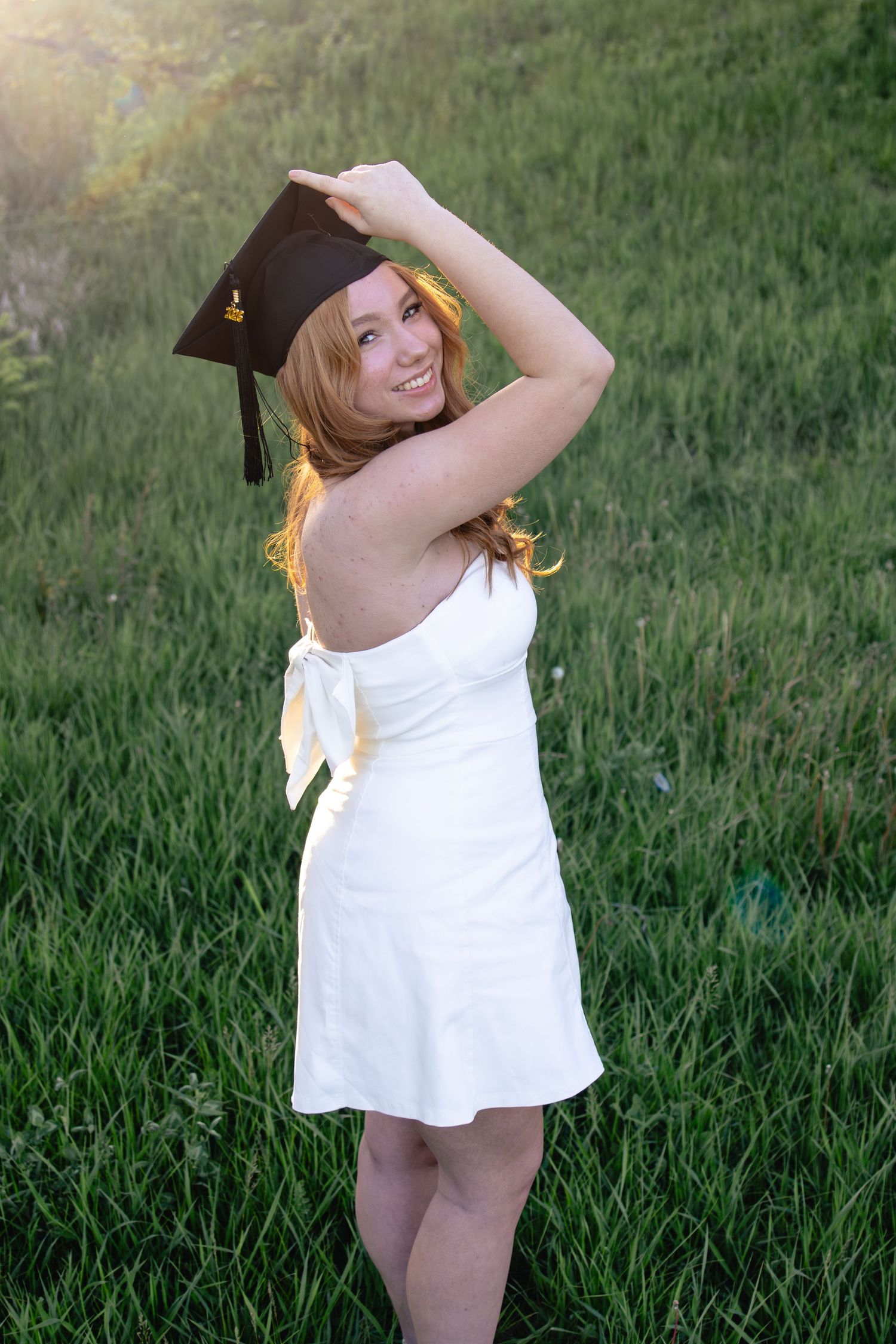 Smiling redhead woman in white dress holding graduation cap outdoors in green field during golden hour.