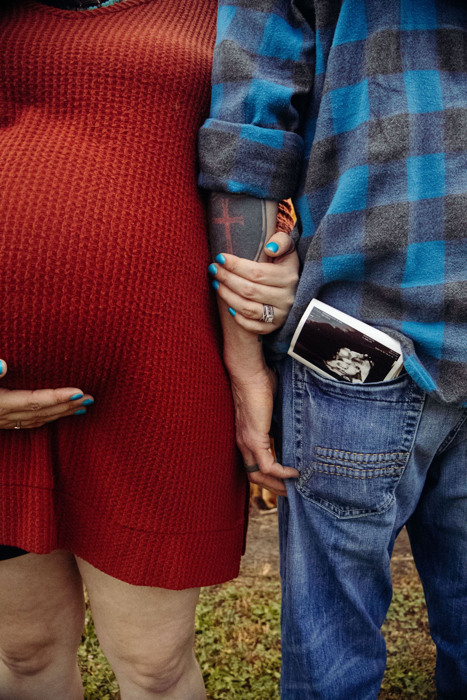 Close-up of couple wearing red sweater and plaid shirt showing ultrasound photo in jean pocket.