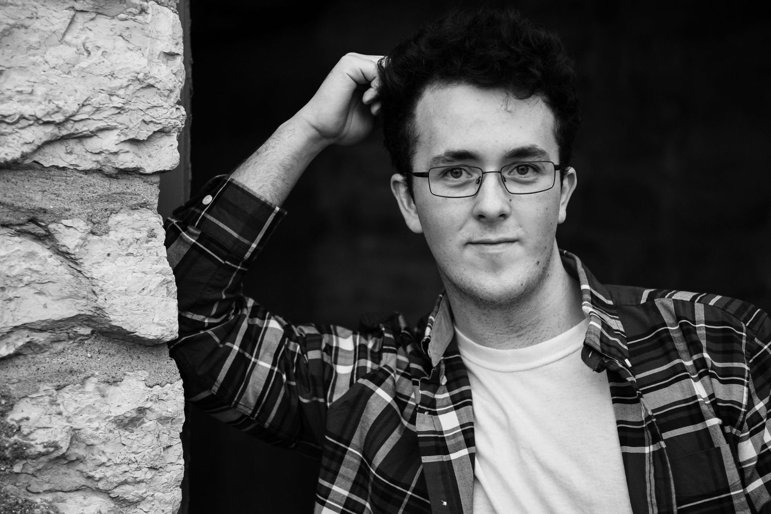 Black and white portrait of person in plaid shirt and glasses against stone wall.