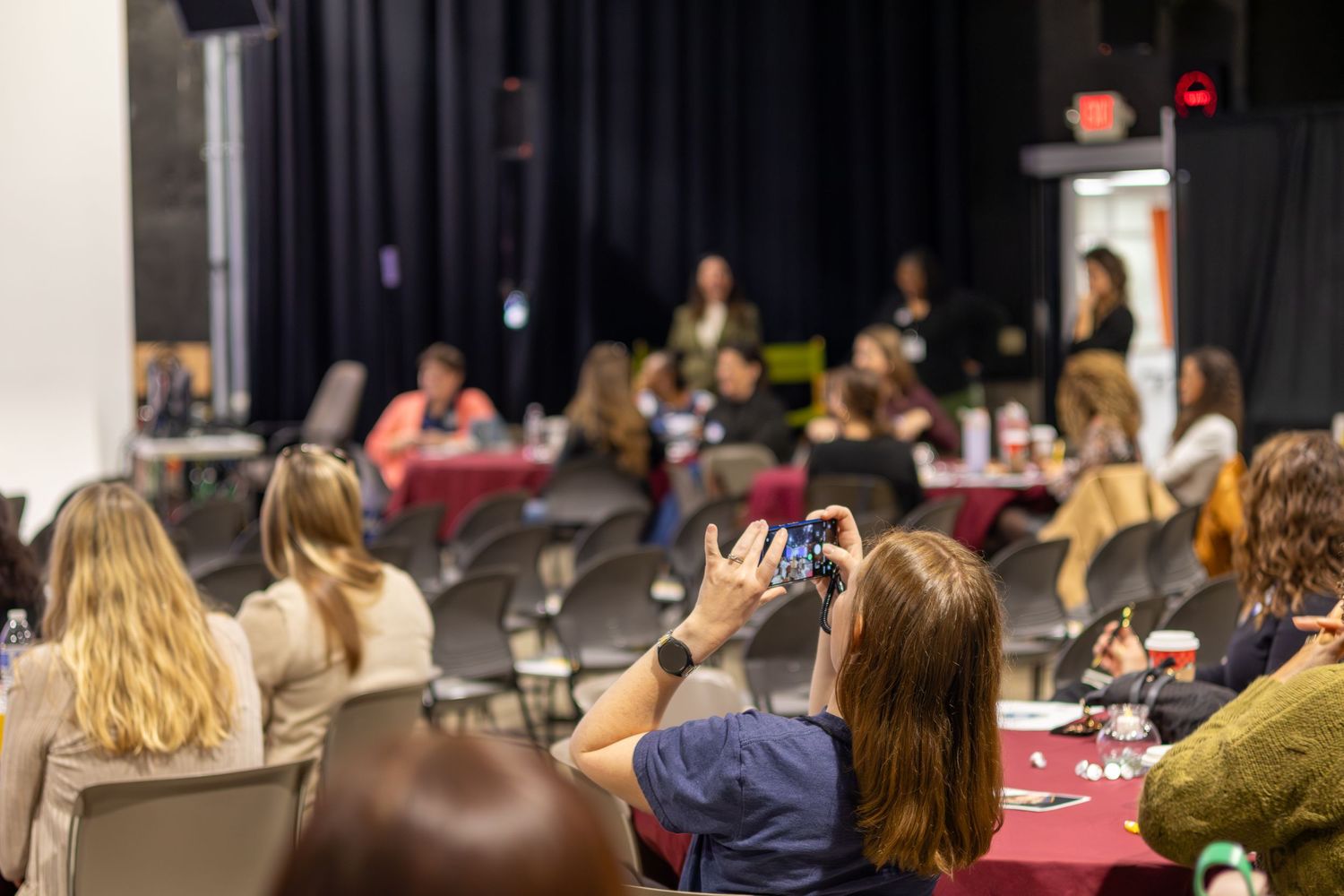 Audience members seated in rows at a conference or presentation venue with dark lighting and red accents.