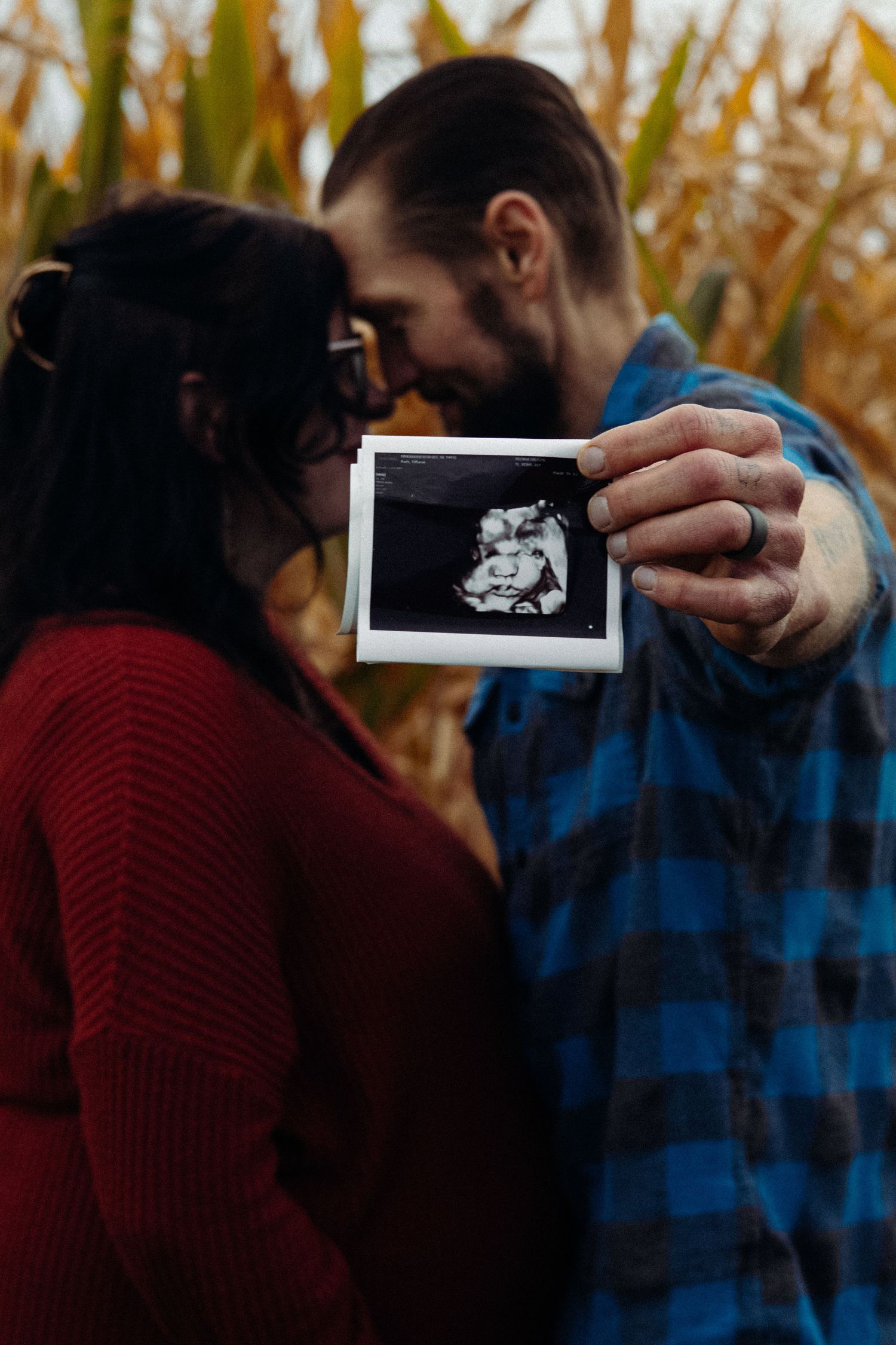 A couple shares an intimate moment while holding up an ultrasound photo in a cornfield.