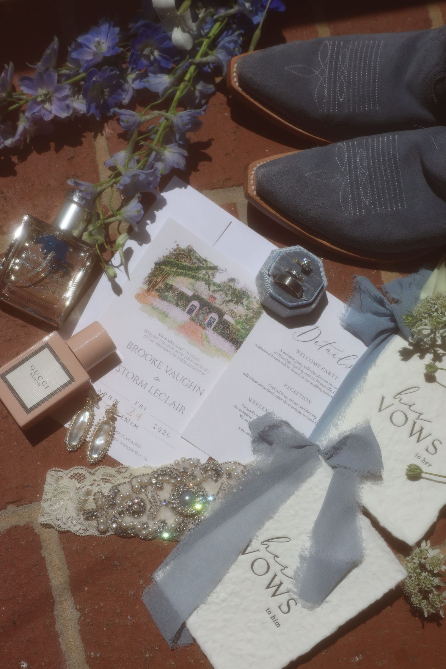 Wedding flat lay with invitations, vow books, perfume, earrings, lace garter, ring box, and gray boots on brick.