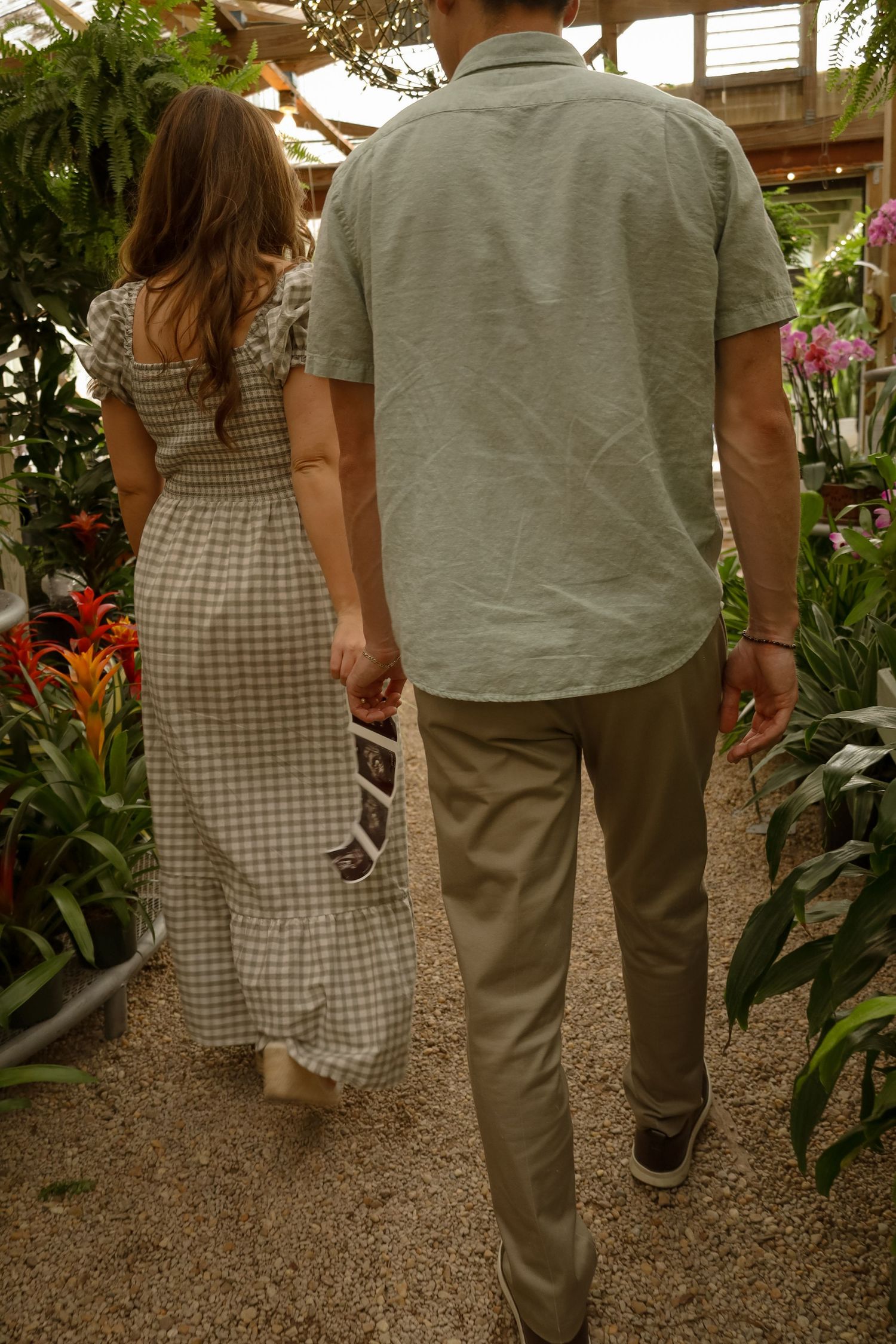 Couple walking hand in hand through a lush greenhouse, wearing neutral tones, surrounded by tropical plants.
