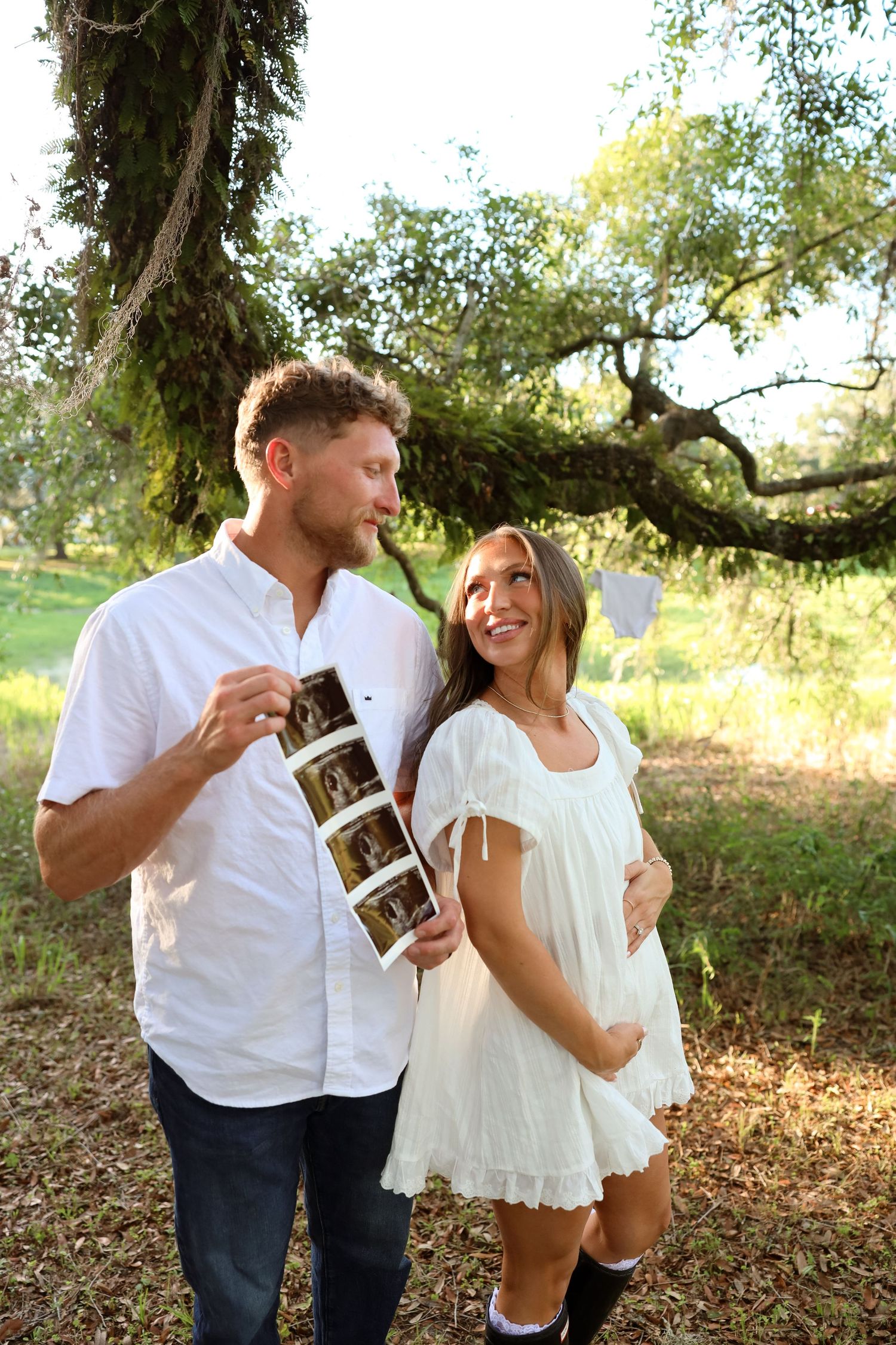 Happy couple outdoors holding ultrasound photos, woman in white dress with baby bump, man in white shirt, under mossy tree.