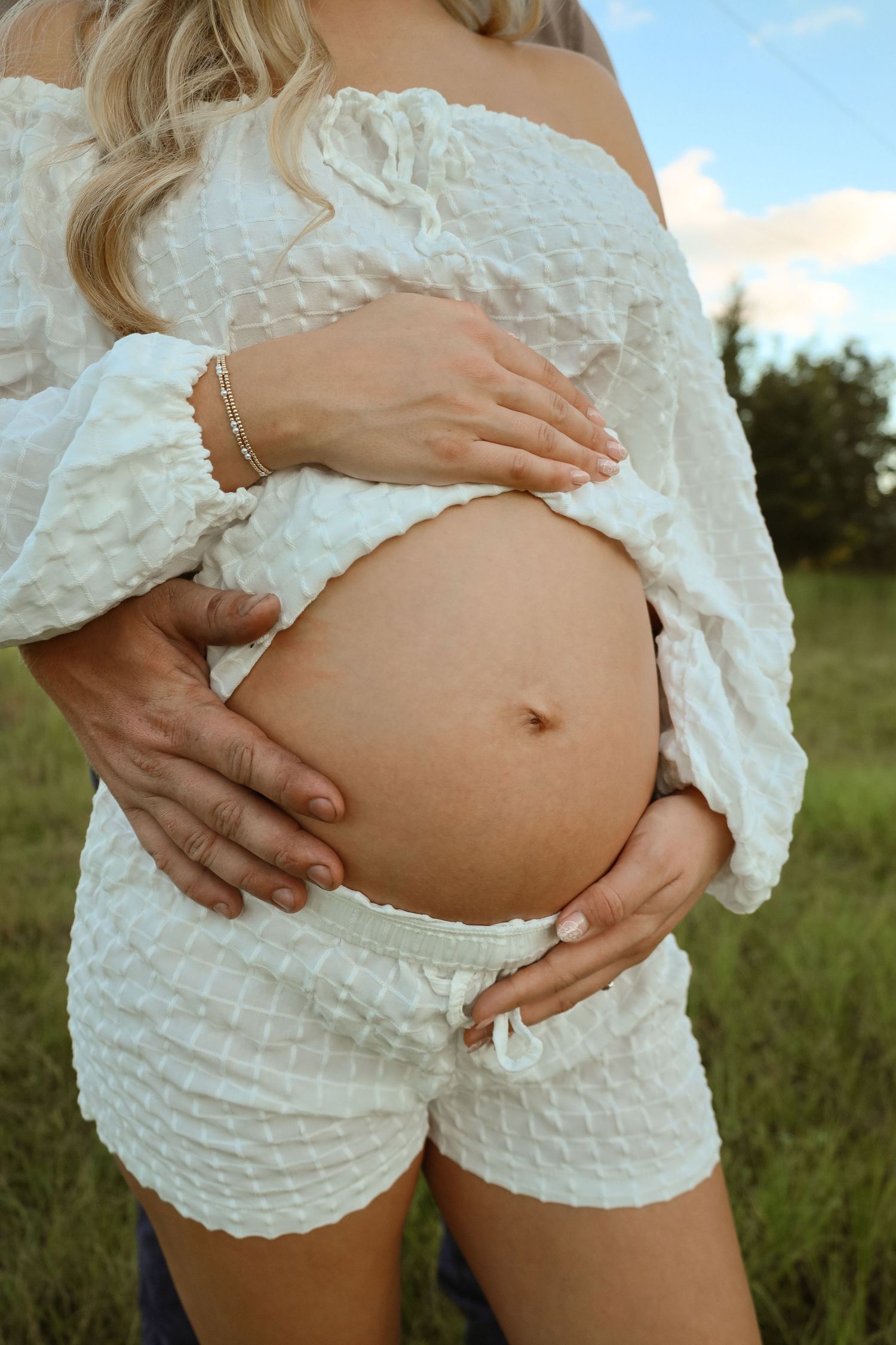 Pregnant woman in a white textured off-shoulder top and shorts cradling her bare baby bump in a grassy field at sunset.