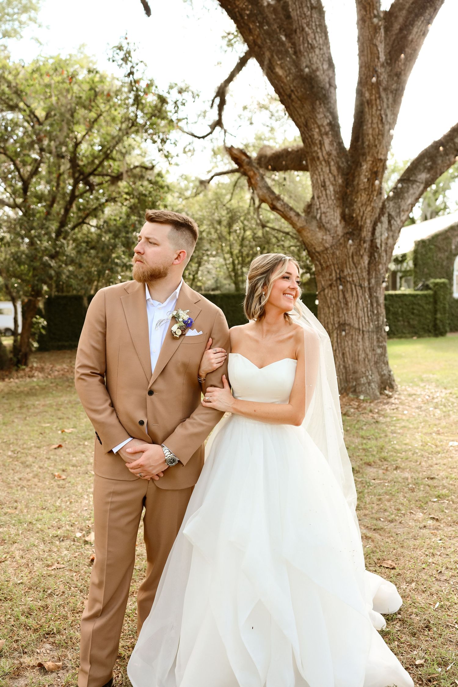 Bride in white strapless ball gown and groom in tan suit pose together outdoors under a large oak tree.