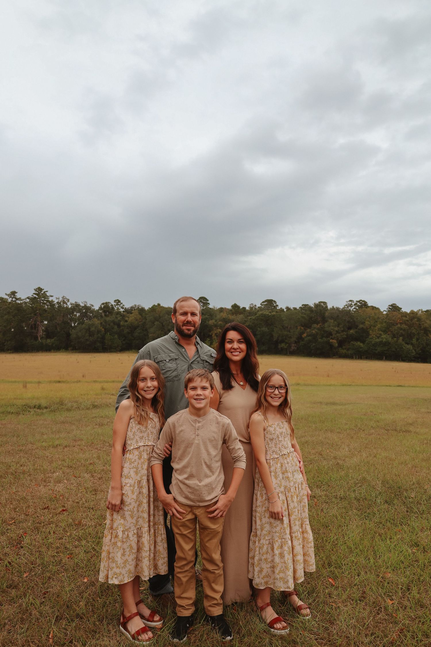 A family stands together in a grassy field on an overcast day wearing coordinated neutral-toned outfits.