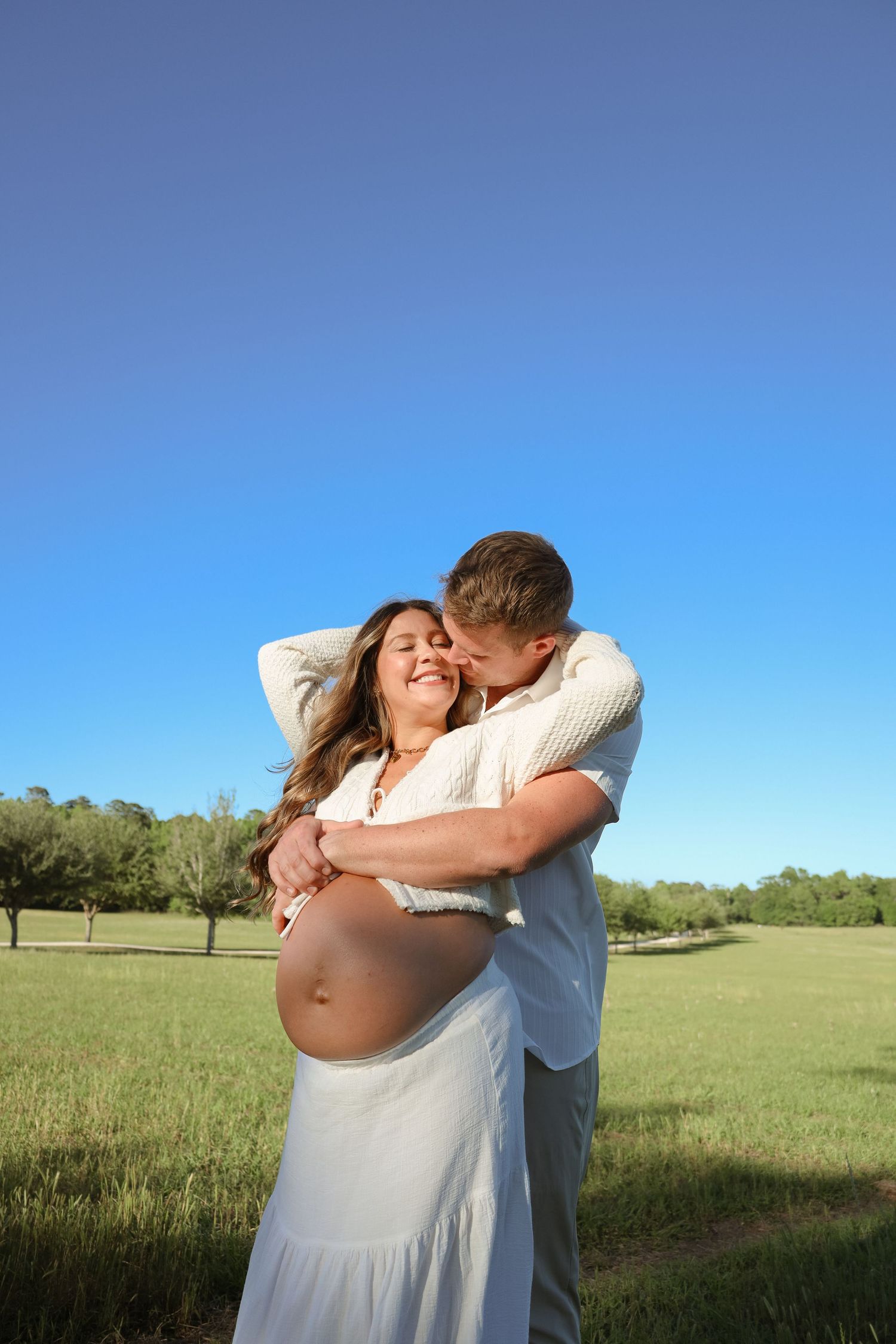 Couple sharing a tender embrace in a sunny green field, with the woman's pregnant belly visible, under a clear blue sky.