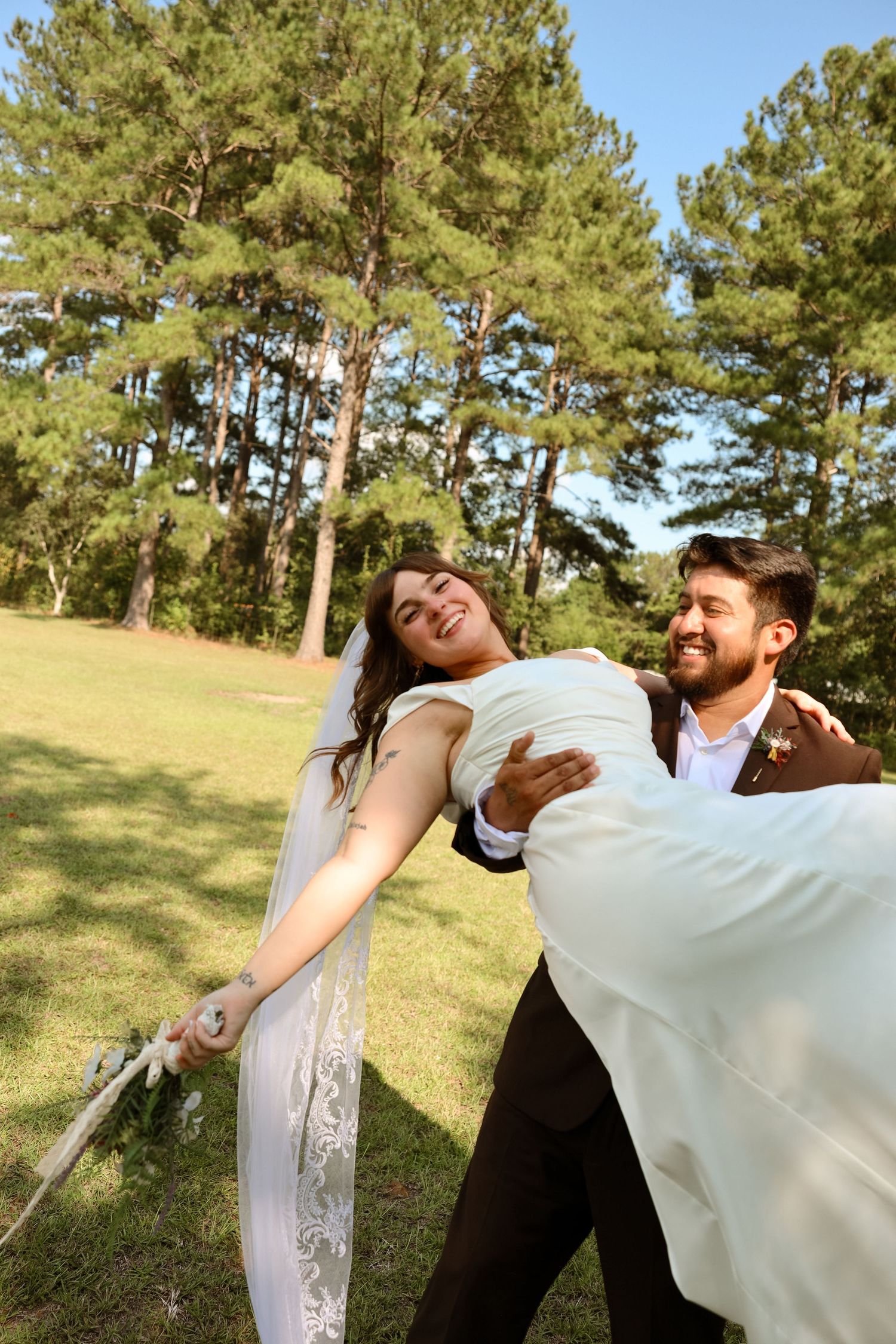 A joyful bride and groom celebrate outdoors in a lush green park, with the groom dipping his smiling bride.