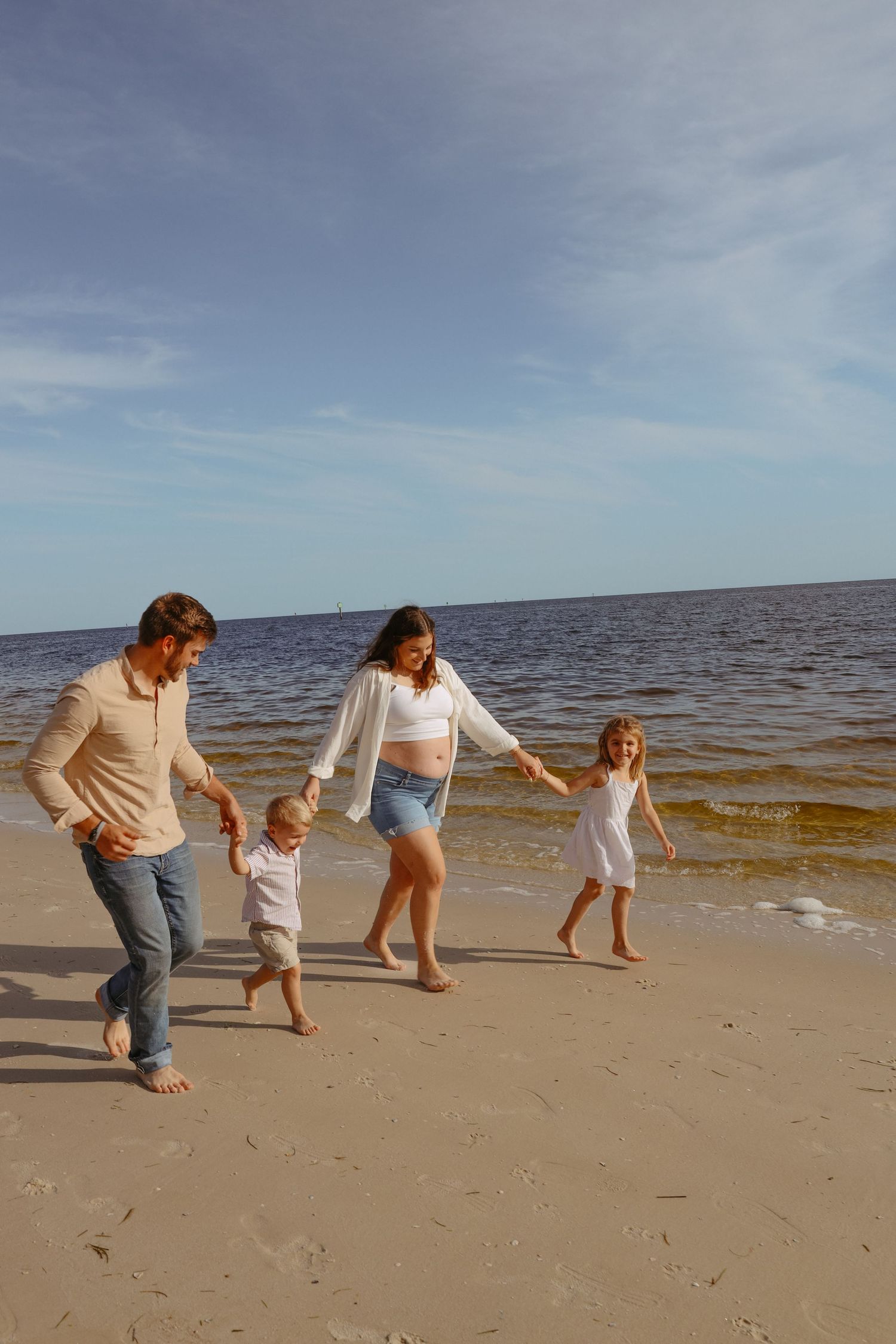 A family plays and runs together along a sandy beach with blue ocean waters and cloudy skies in the background.