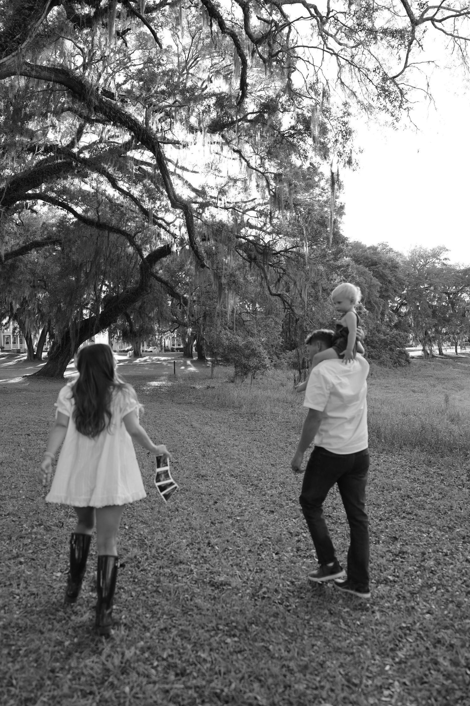 Black and white photo of a family walking in a park, father carrying child on shoulders, daughter in white dress and boots.