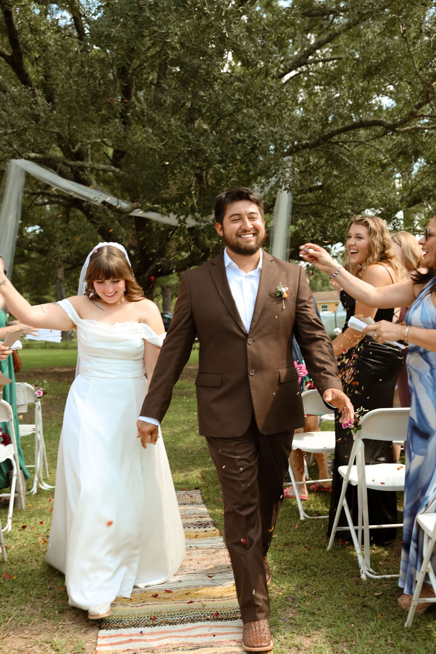 Smiling newlywed couple walks down the aisle at an outdoor wedding ceremony as guests toss confetti.