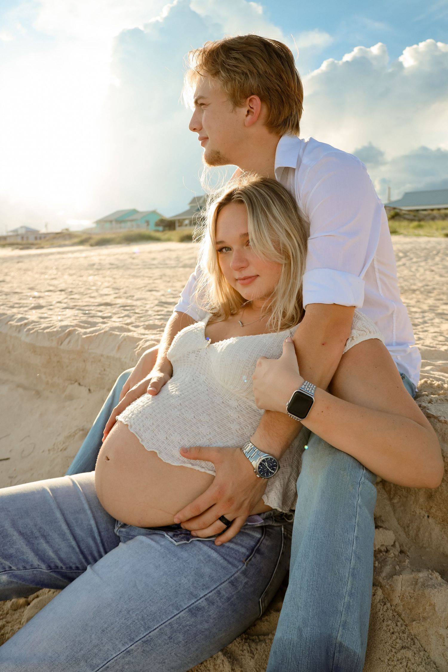 Pregnant couple sitting on beach sand, man in white shirt embracing woman holding baby bump, sunny sky backdrop.