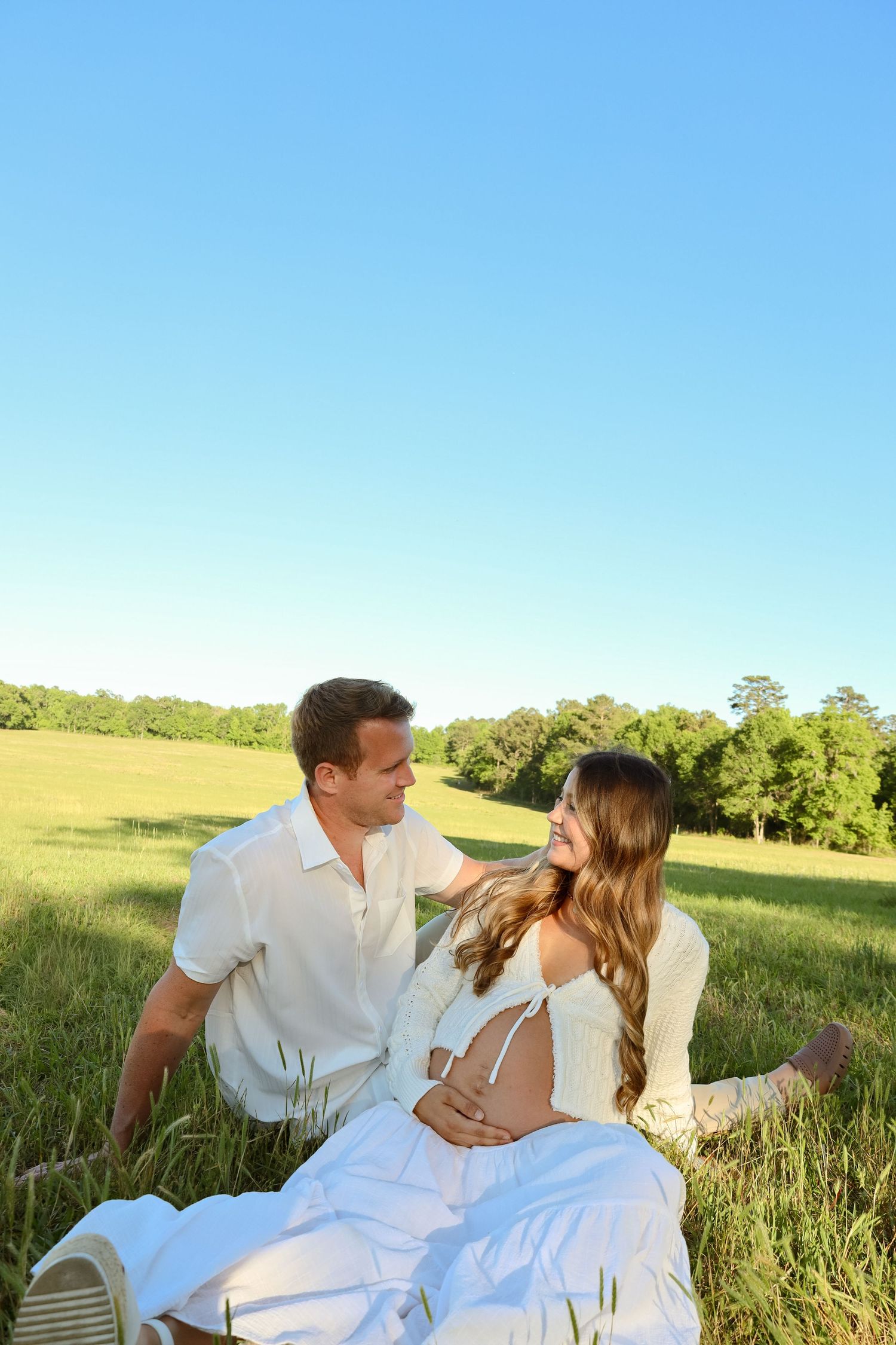 Happy pregnant couple sitting in a green field on a sunny day, smiling at each other in white outfits.