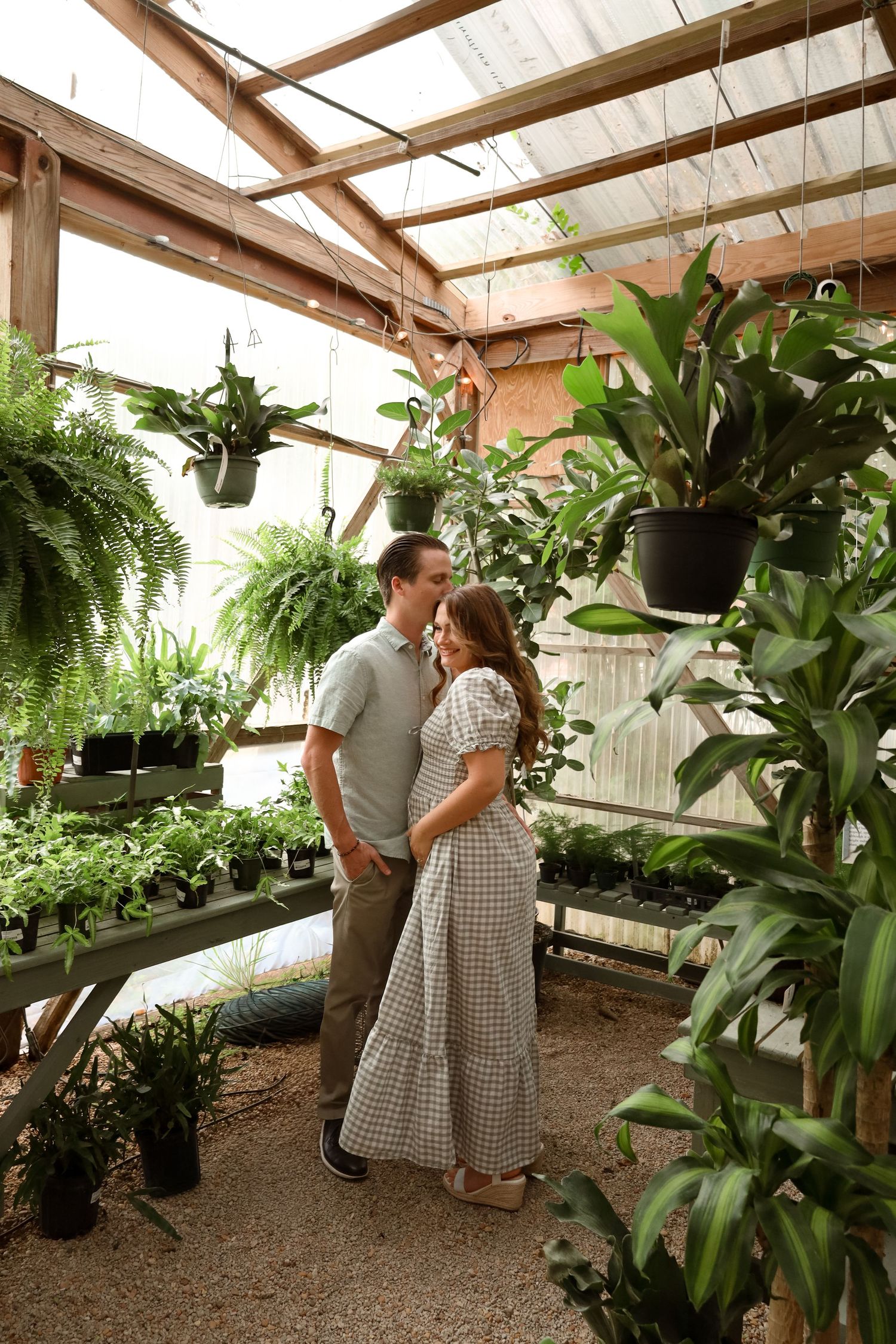 Couple sharing a romantic moment surrounded by lush green plants in a sunlit greenhouse.