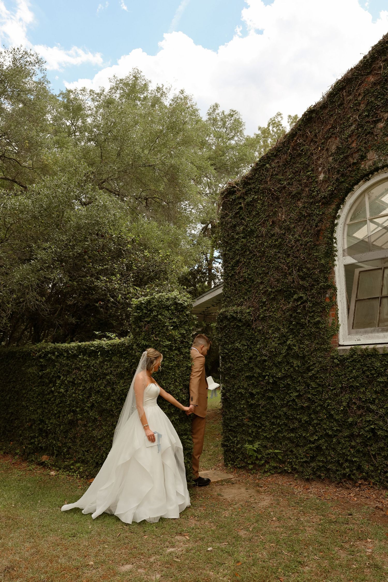 Bride and groom share a first look moment beside an ivy-covered building surrounded by lush greenery.