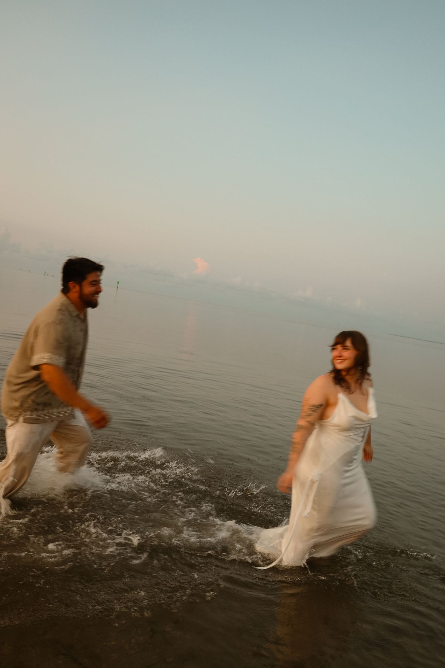 A couple joyfully splashing through shallow ocean waves at sunset, woman in white dress and man in casual attire.