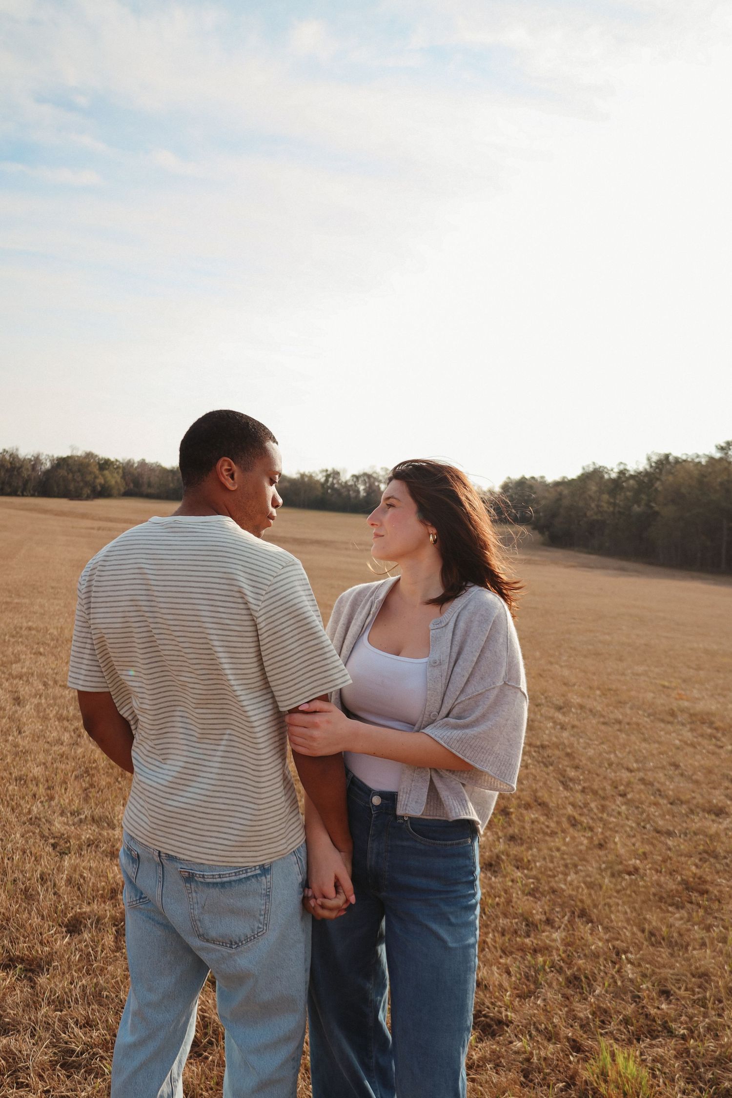 A couple stands together holding hands in a golden wheat field at sunset.