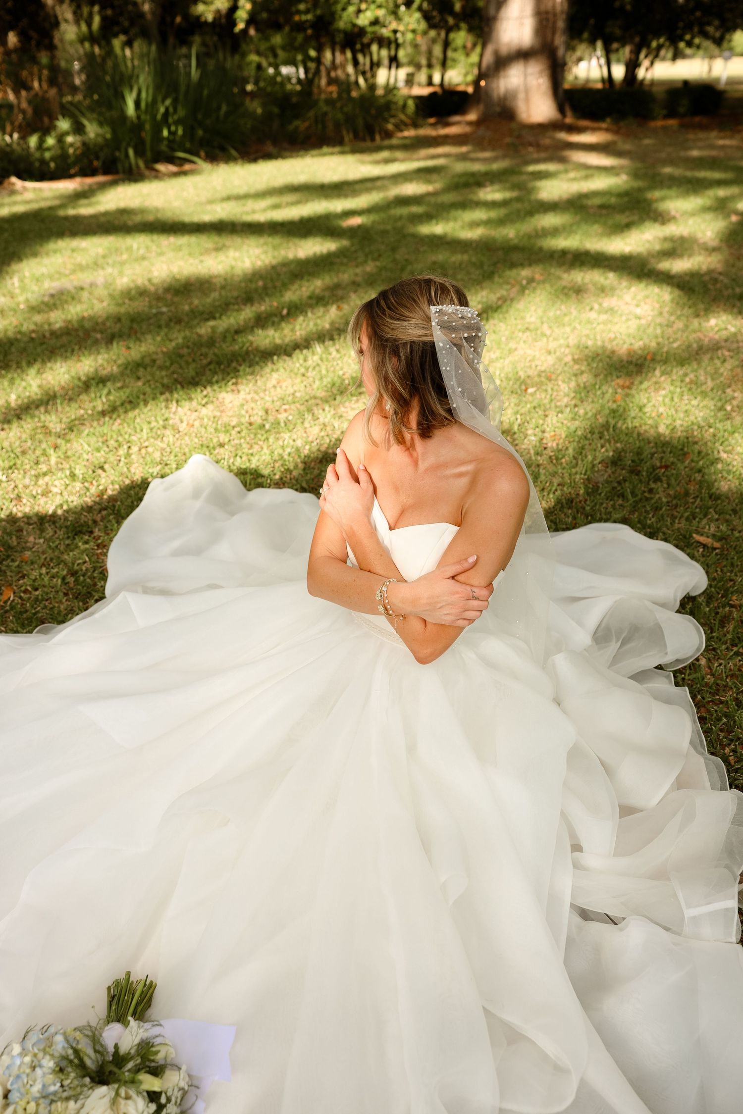 Bride in a flowing white ball gown and veil sits on green grass, viewed from behind, with a bouquet nearby.