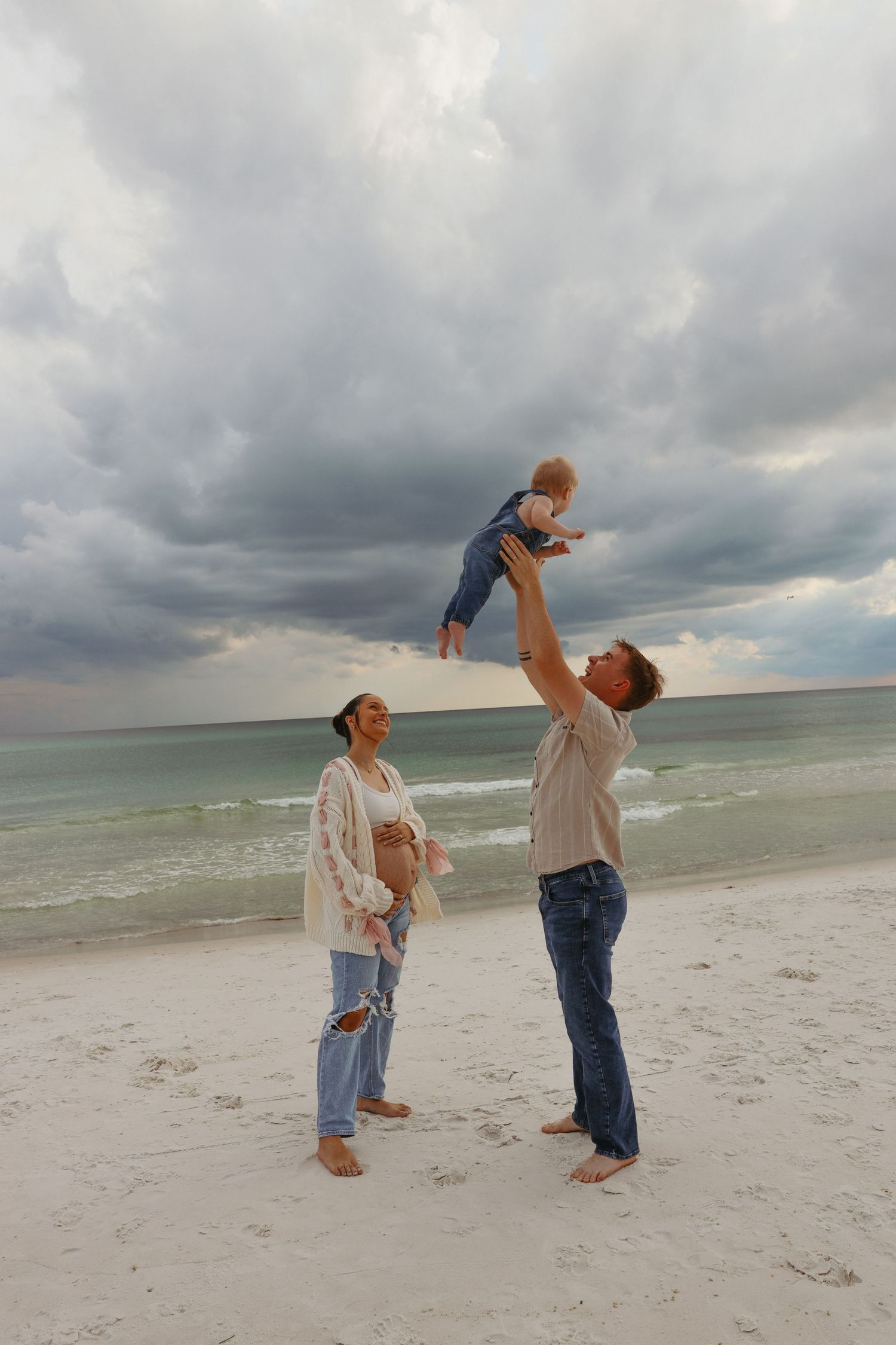 Two people standing on a white sandy beach under dramatic storm clouds lifting a small child into the air.
