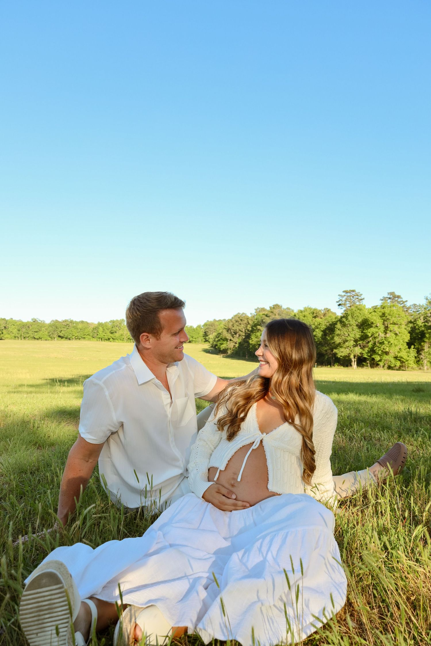 A happy couple sits in a green meadow, the pregnant woman in white cradling her belly while her partner gazes at her lovingly.
