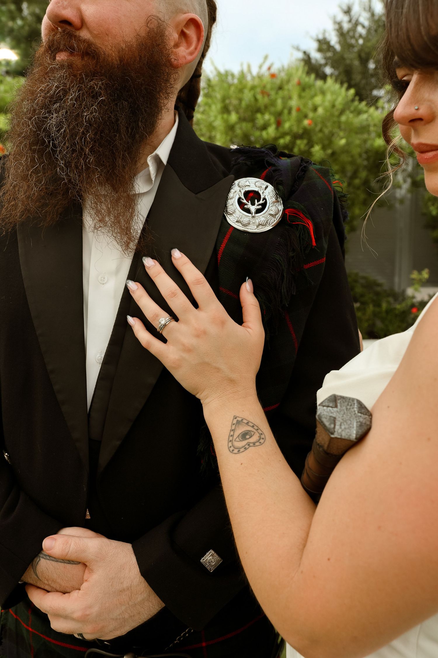 Bride with heart tattoo on wrist places hand on groom's black tuxedo jacket with Scottish thistle brooch at wedding.