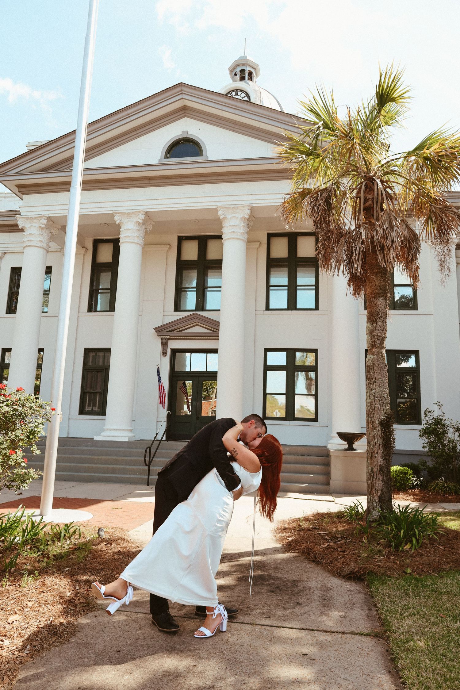 A romantic couple shares a dip-style dance pose in front of a white colonial building with palm trees.