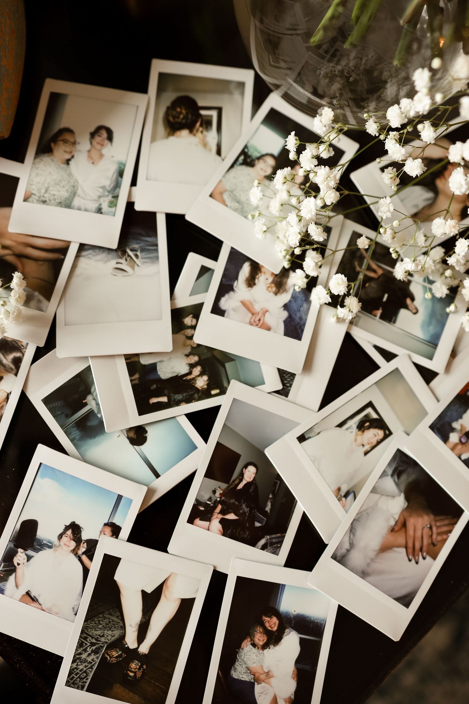 A collection of Polaroid photos scattered on a dark table, featuring candid moments, alongside delicate white baby's breath flowers.